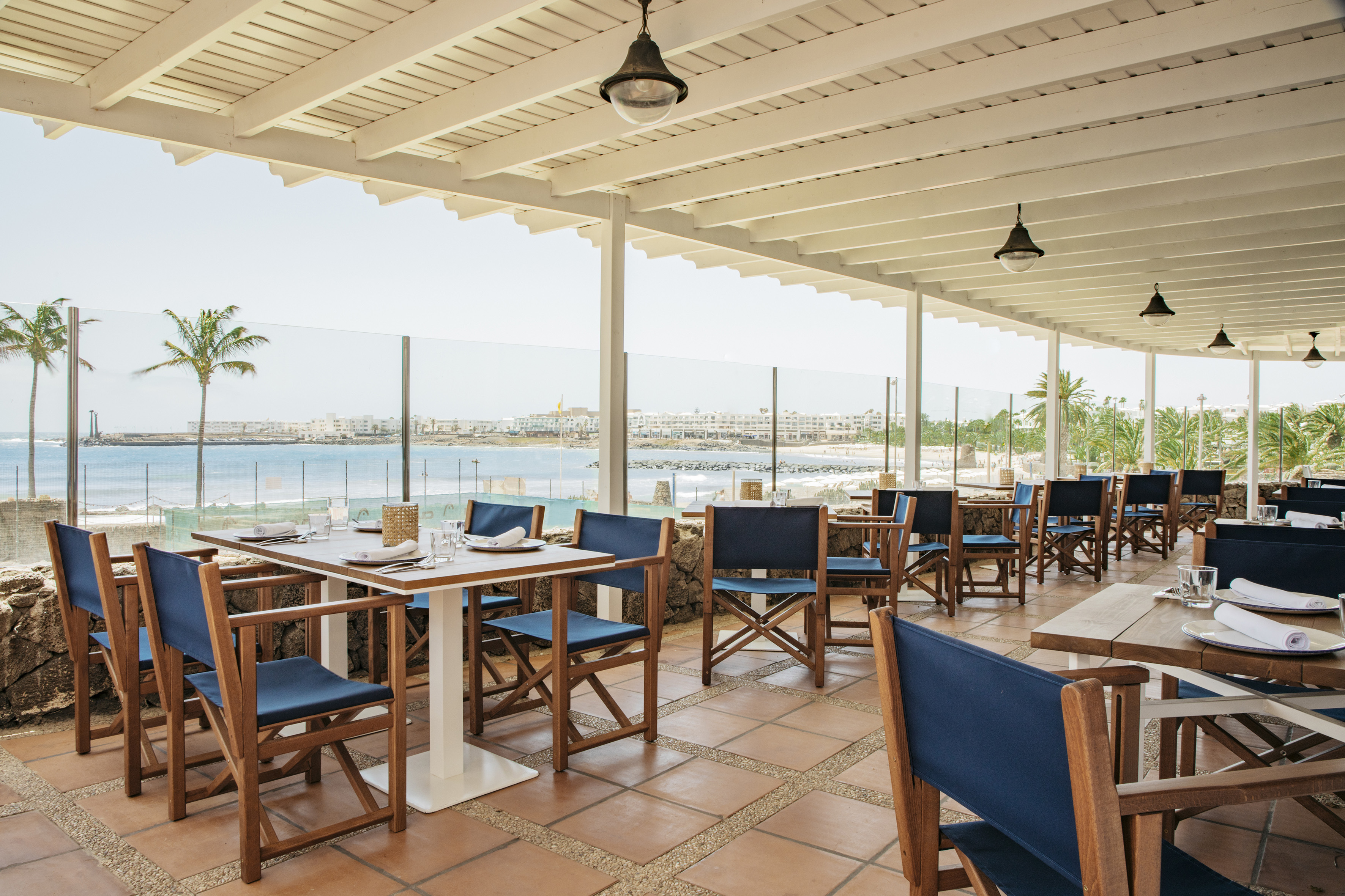 Outdoor restaurant patio with ocean view, blue chairs, and palm trees.