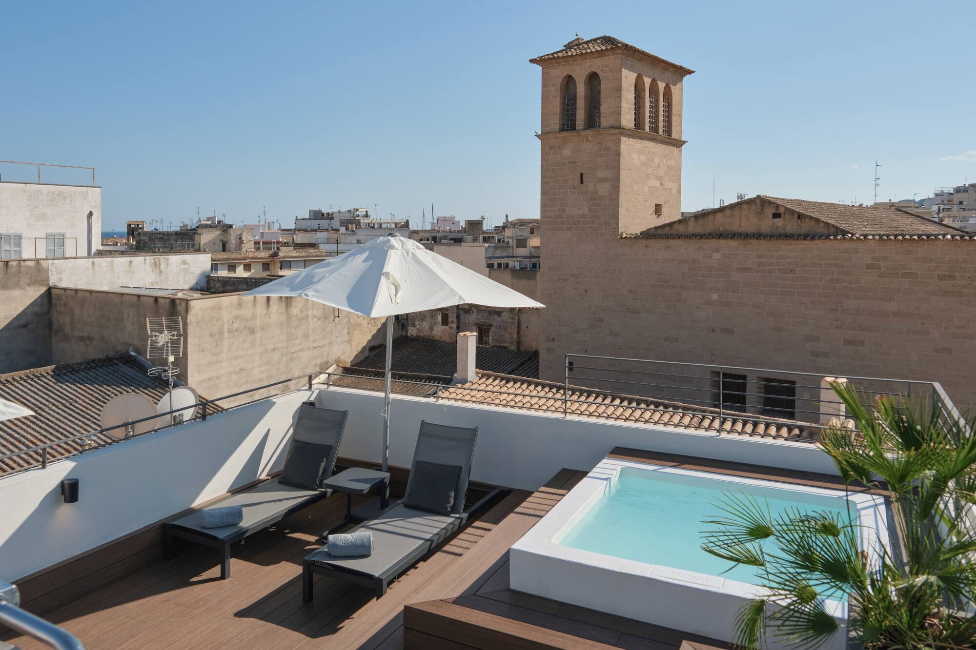 Rooftop terrace with hot tub, loungers, umbrella, and historic city tower view.
