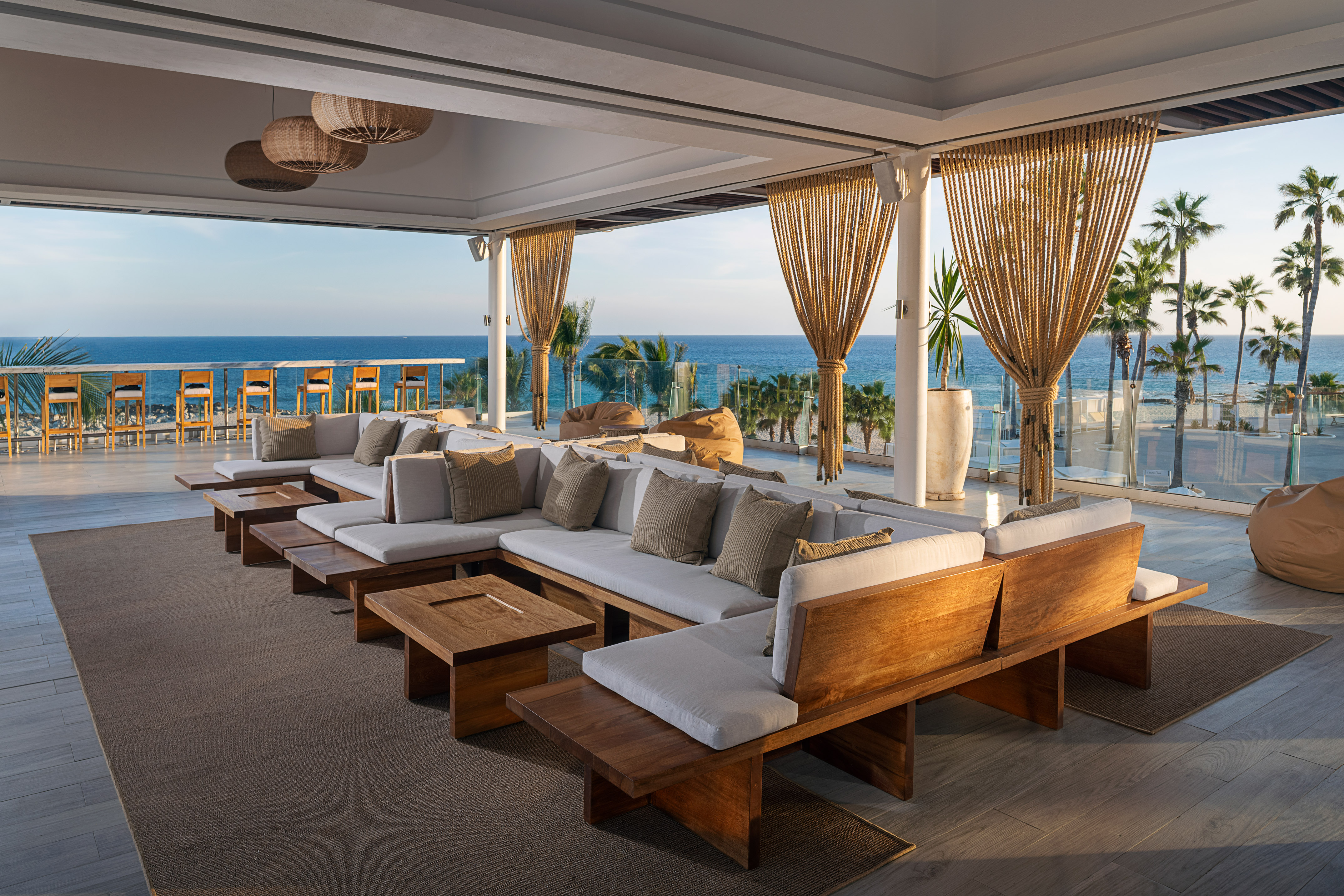 Outdoor lounge patio with ocean view, wooden sofas, palm trees, and bar area.