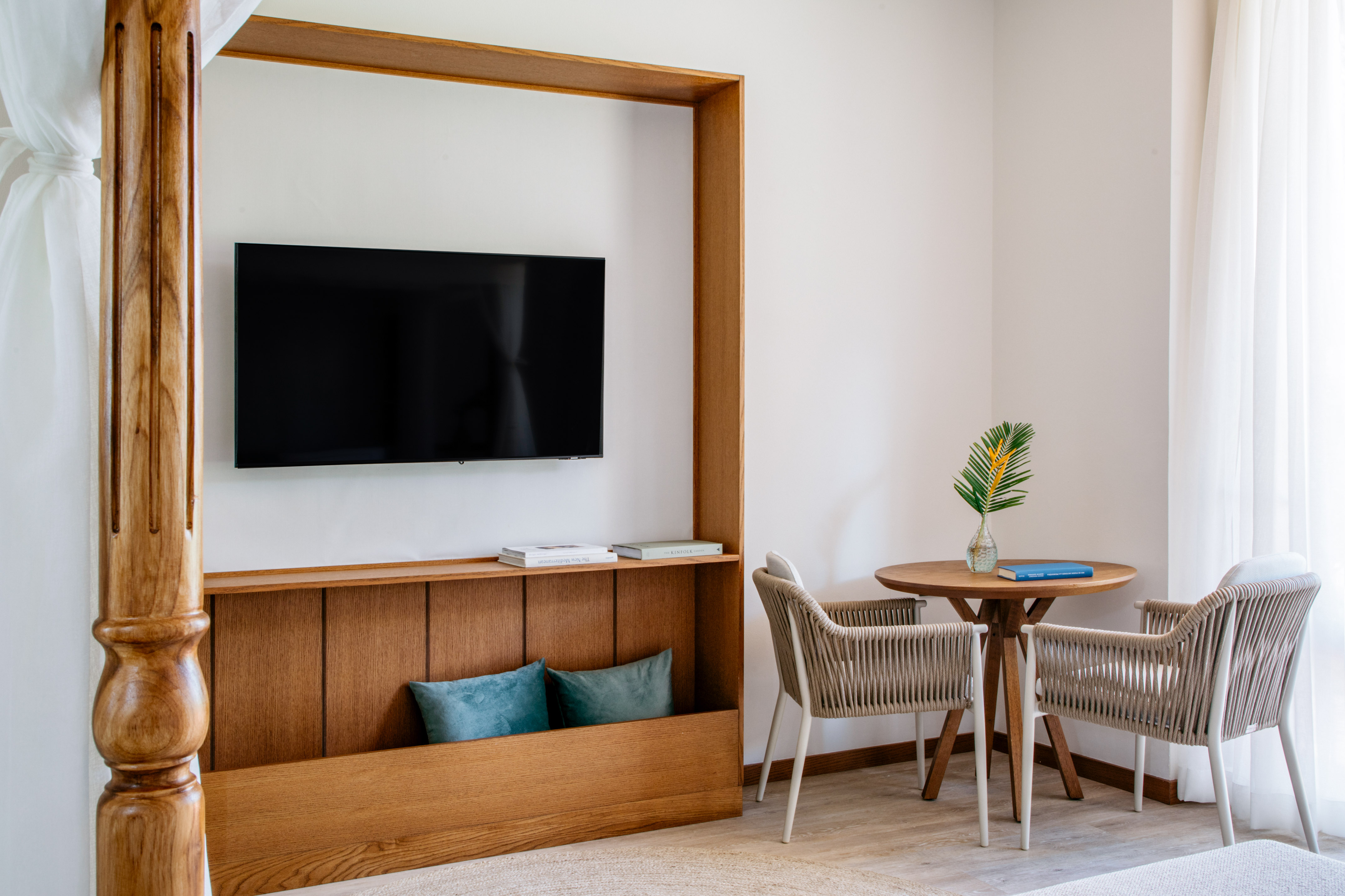 Modern hotel bedroom with wooden four-poster bed, wall TV, and small seating area.
