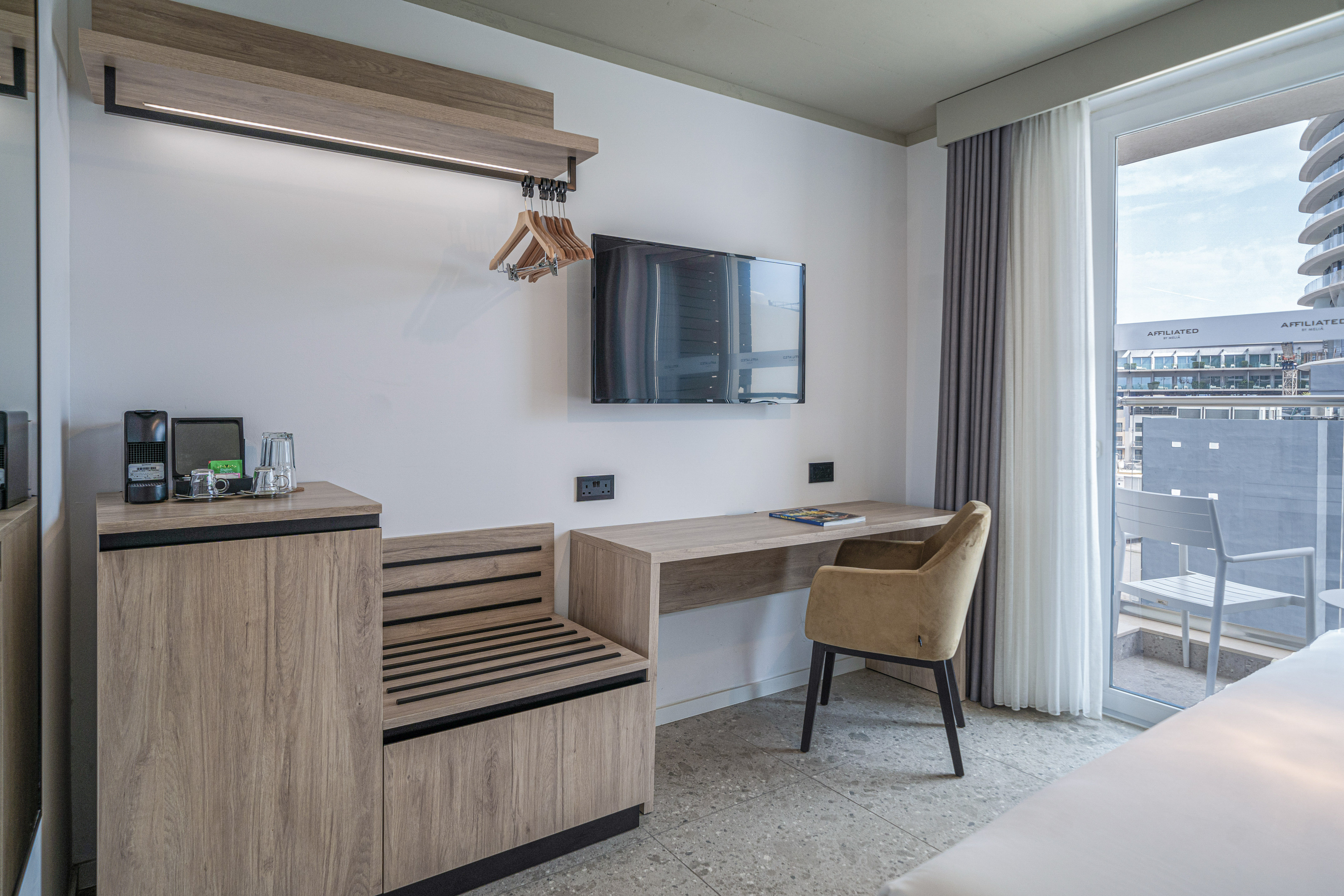 Hotel room with modern desk, wall-mounted TV, coffee corner, and balcony view.