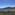 Clear blue bay, green forested hills, rocky coastline, and coastal buildings.