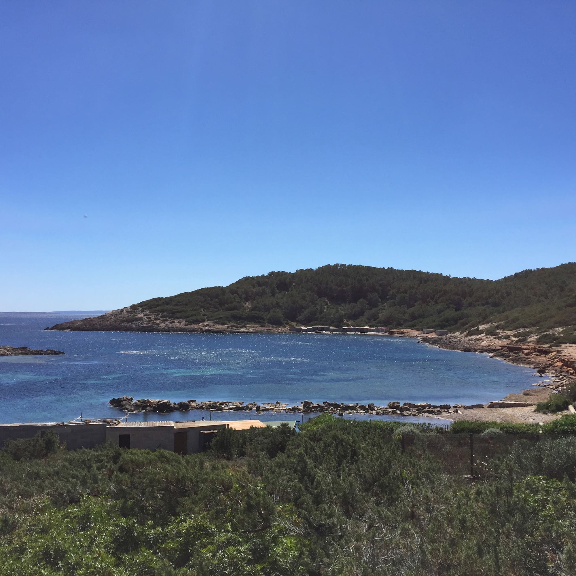 Clear blue bay, green forested hills, rocky coastline, and coastal buildings.