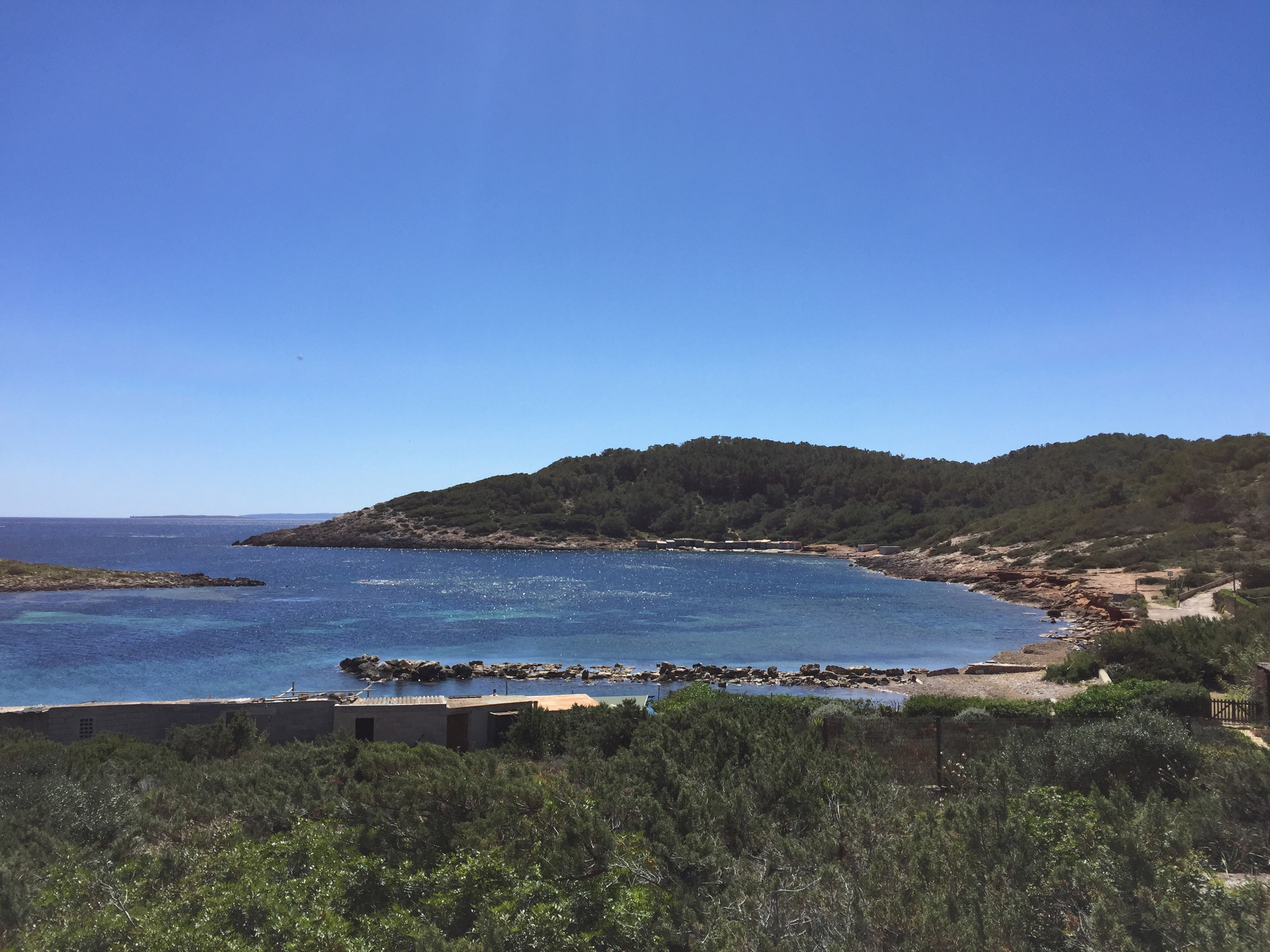 Clear blue bay, green forested hills, rocky coastline, and coastal buildings.