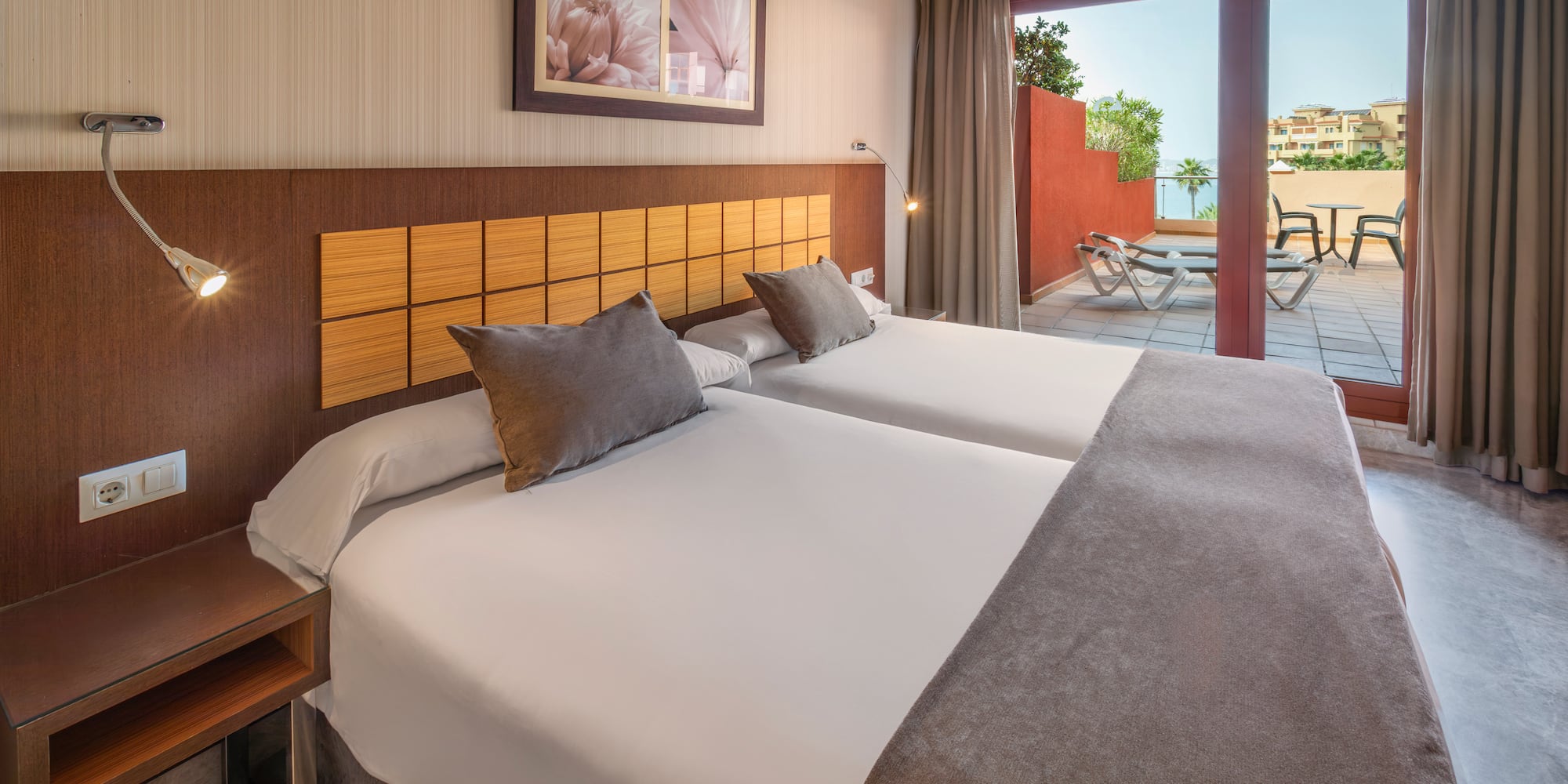 Hotel bedroom with twin beds, private terrace, lounge chairs, and sea view.