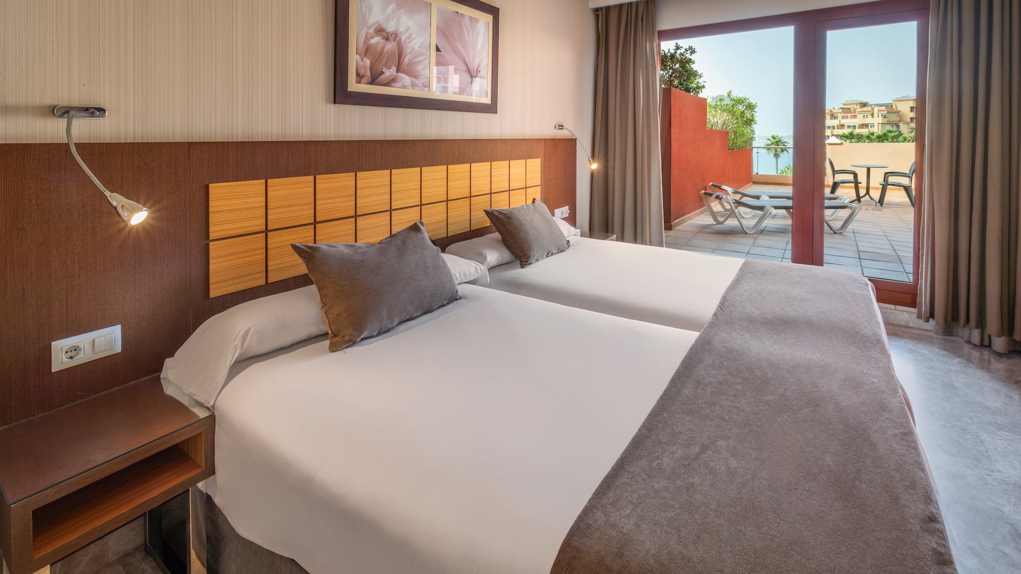 Hotel bedroom with twin beds, private terrace, lounge chairs, and sea view.