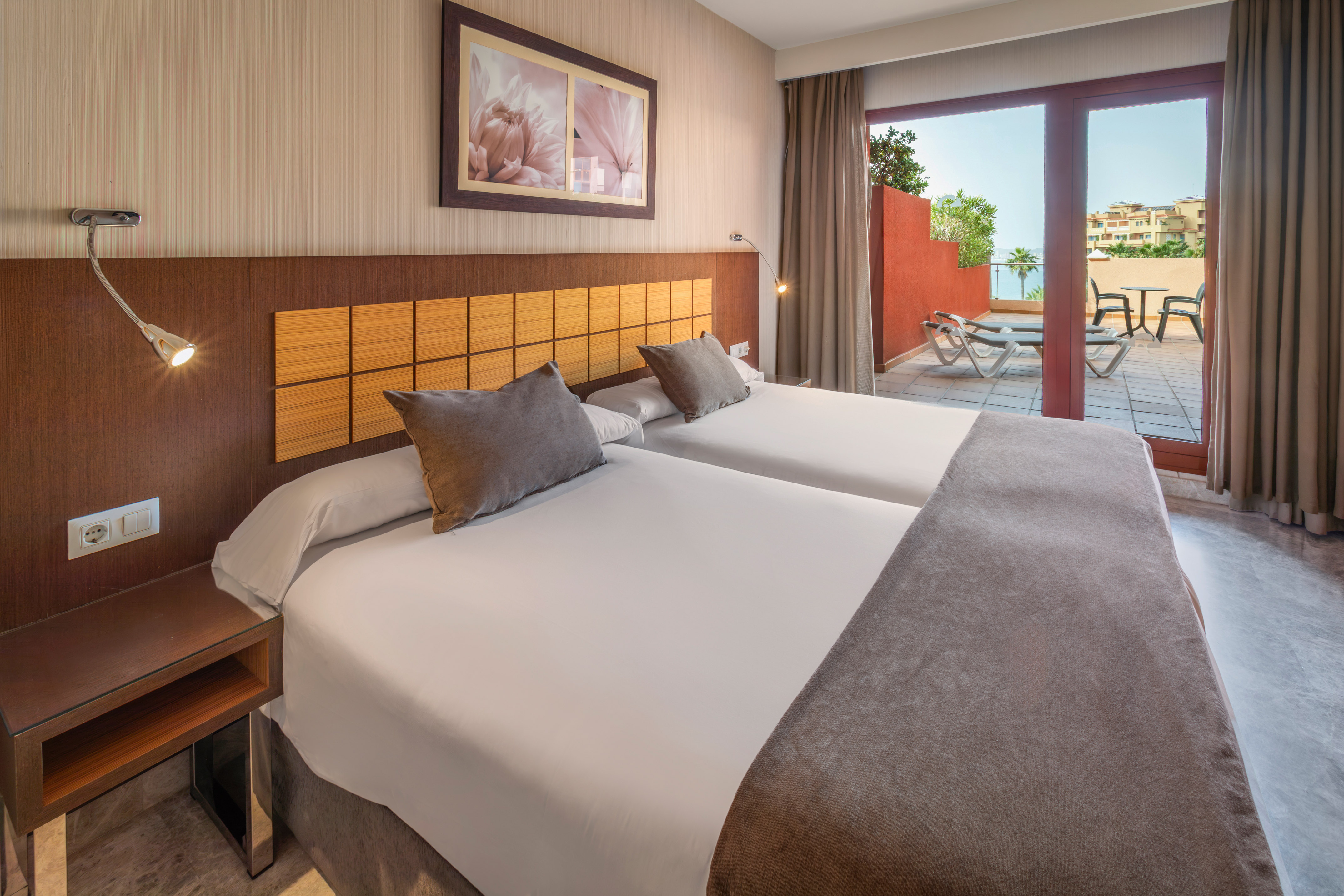Hotel bedroom with twin beds, private terrace, lounge chairs, and sea view.