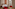 Contemporary hotel room, red chairs, patterned headboard art, city view.