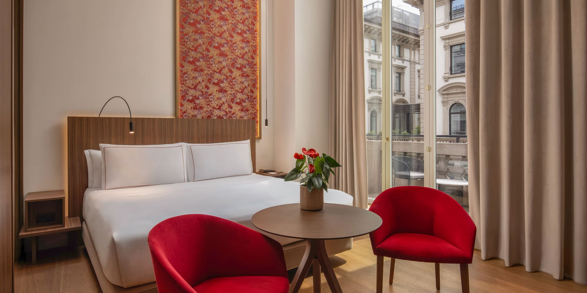 Contemporary hotel room, red chairs, patterned headboard art, city view.