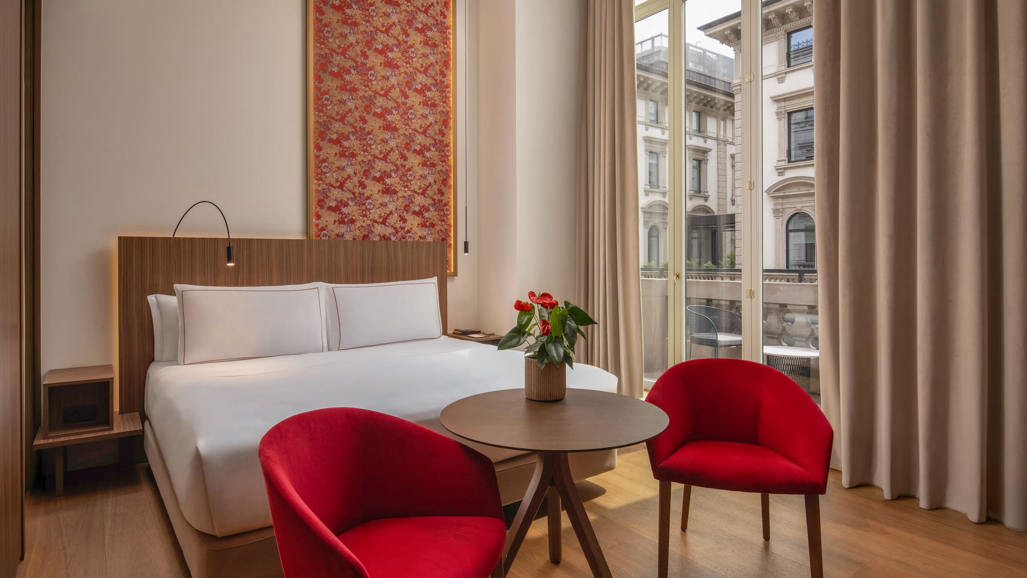 Contemporary hotel room, red chairs, patterned headboard art, city view.