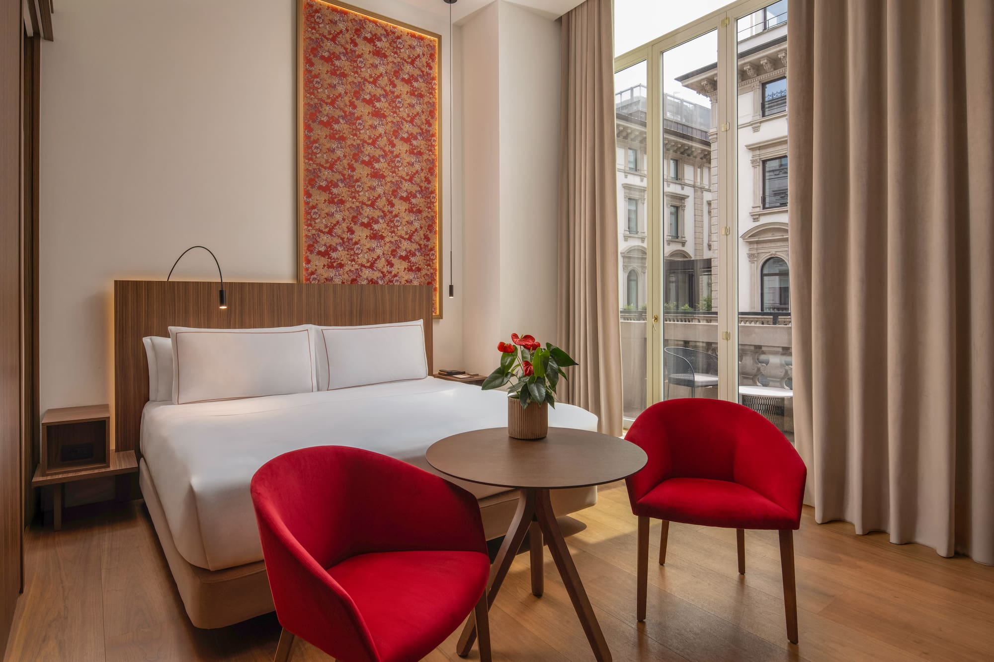 Contemporary hotel room, red chairs, patterned headboard art, city view.