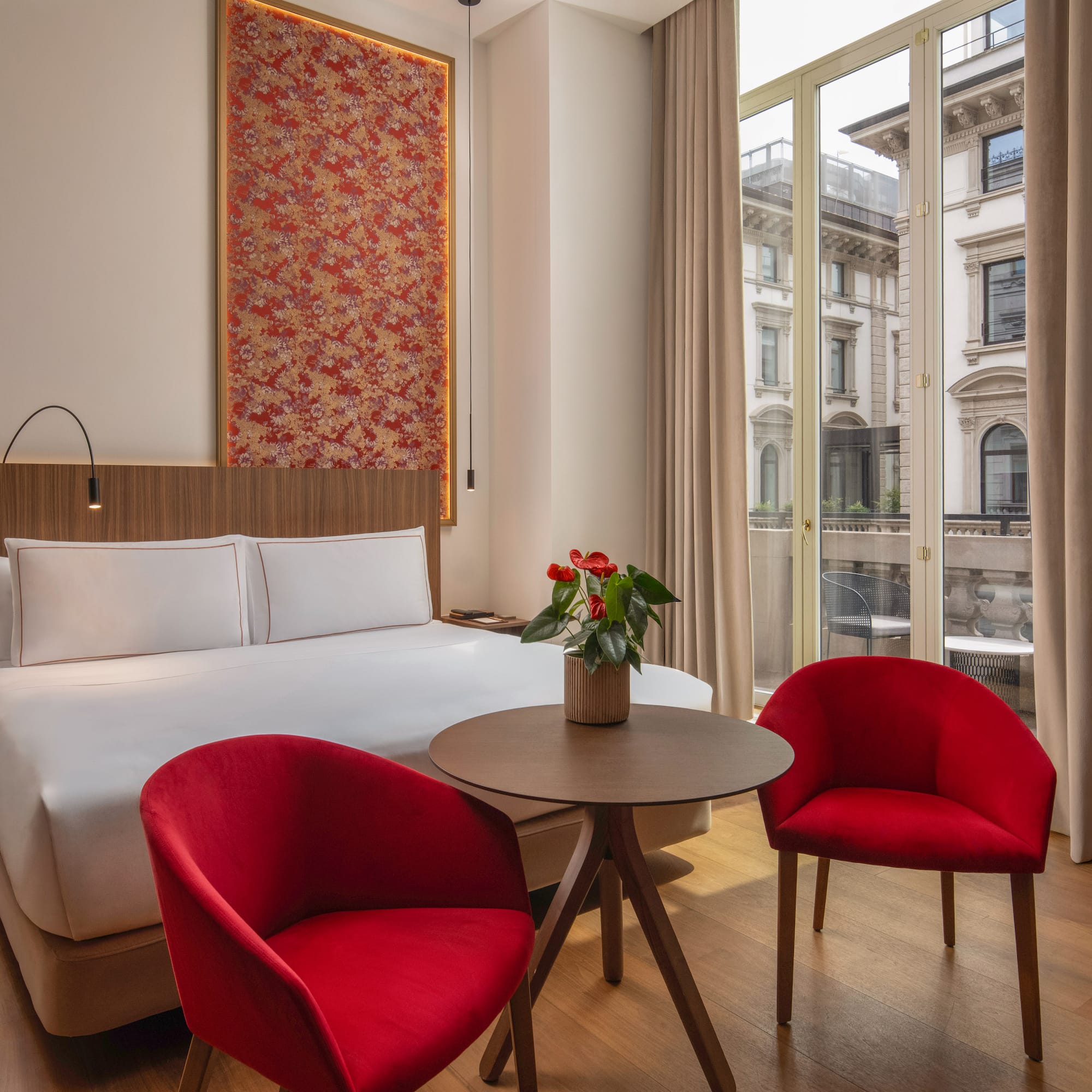 Contemporary hotel room, red chairs, patterned headboard art, city view.
