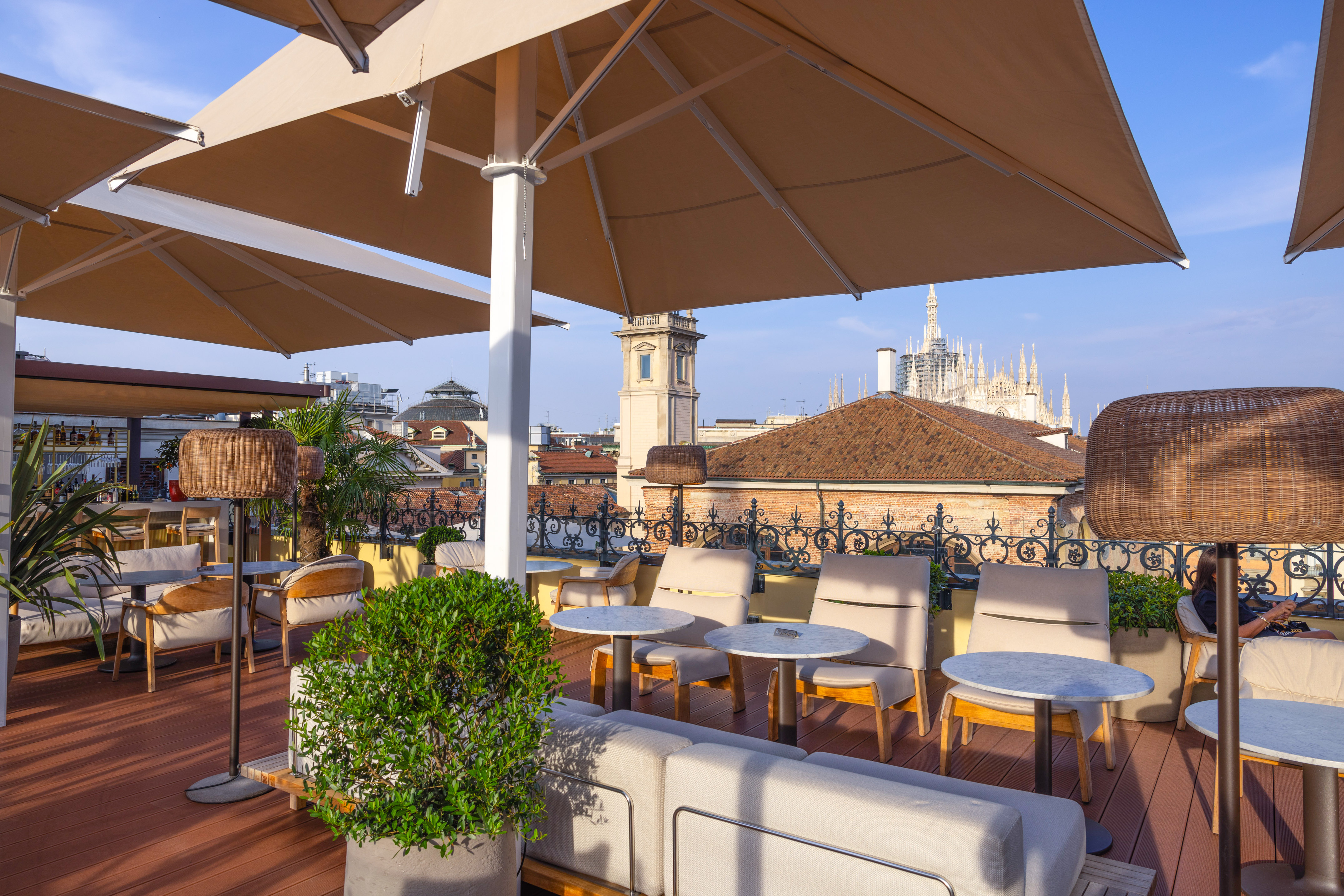 Milan rooftop terrace bar with outdoor seating, brown umbrellas, and Duomo view.