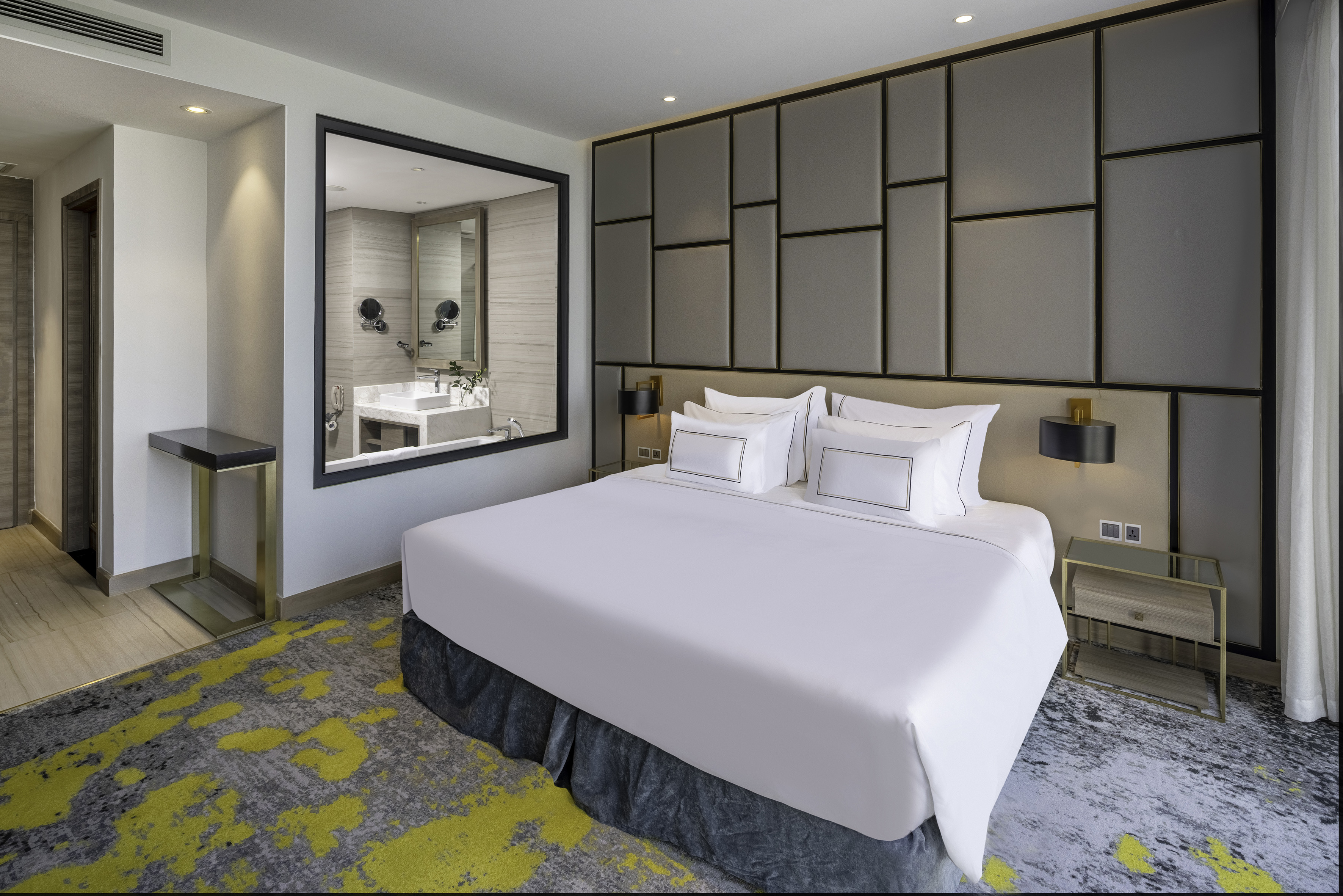 Modern hotel bedroom with king bed, geometric headboard, and grey-yellow carpet.