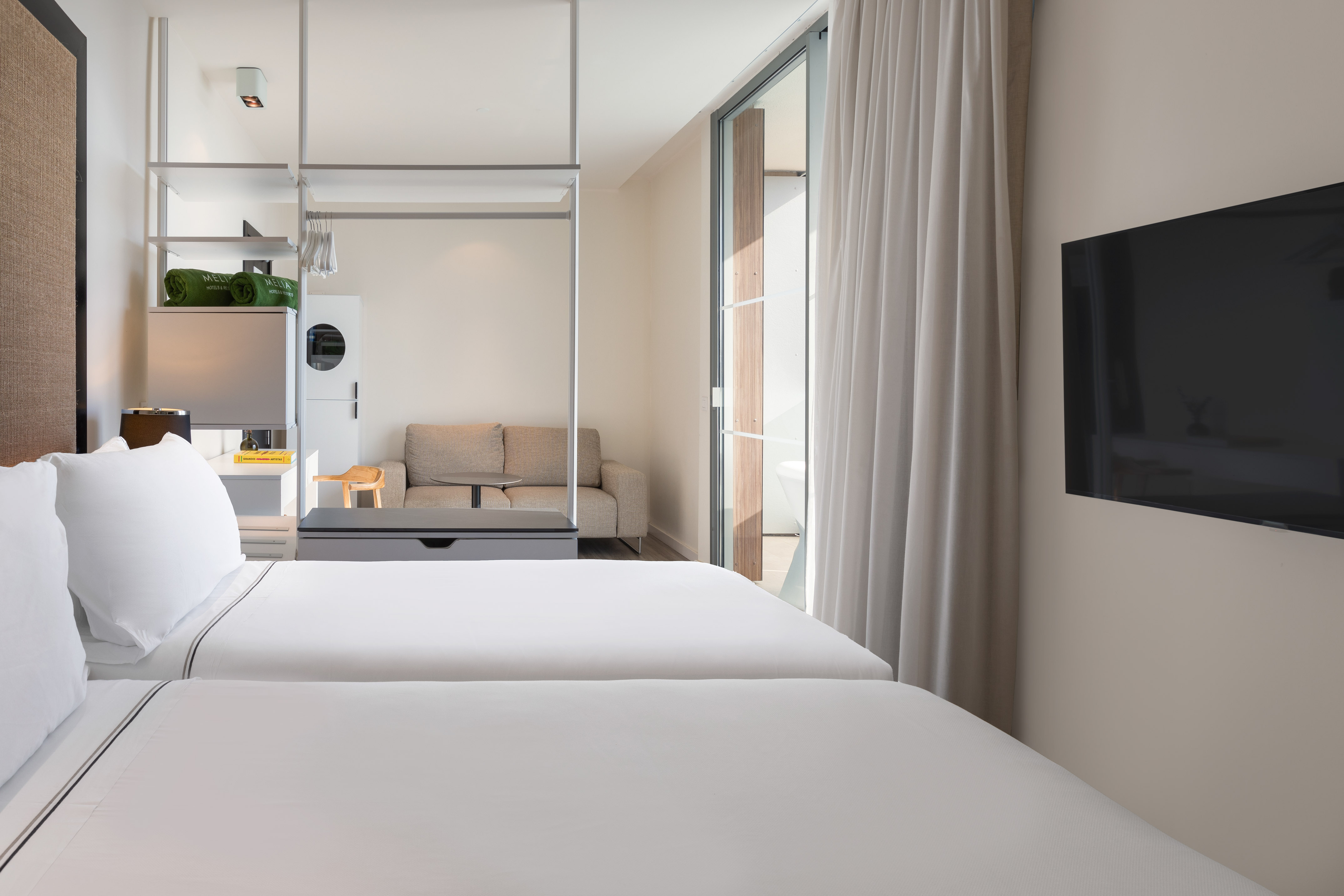Modern minimalist hotel room with twin beds, balcony, wall TV, and sofa.