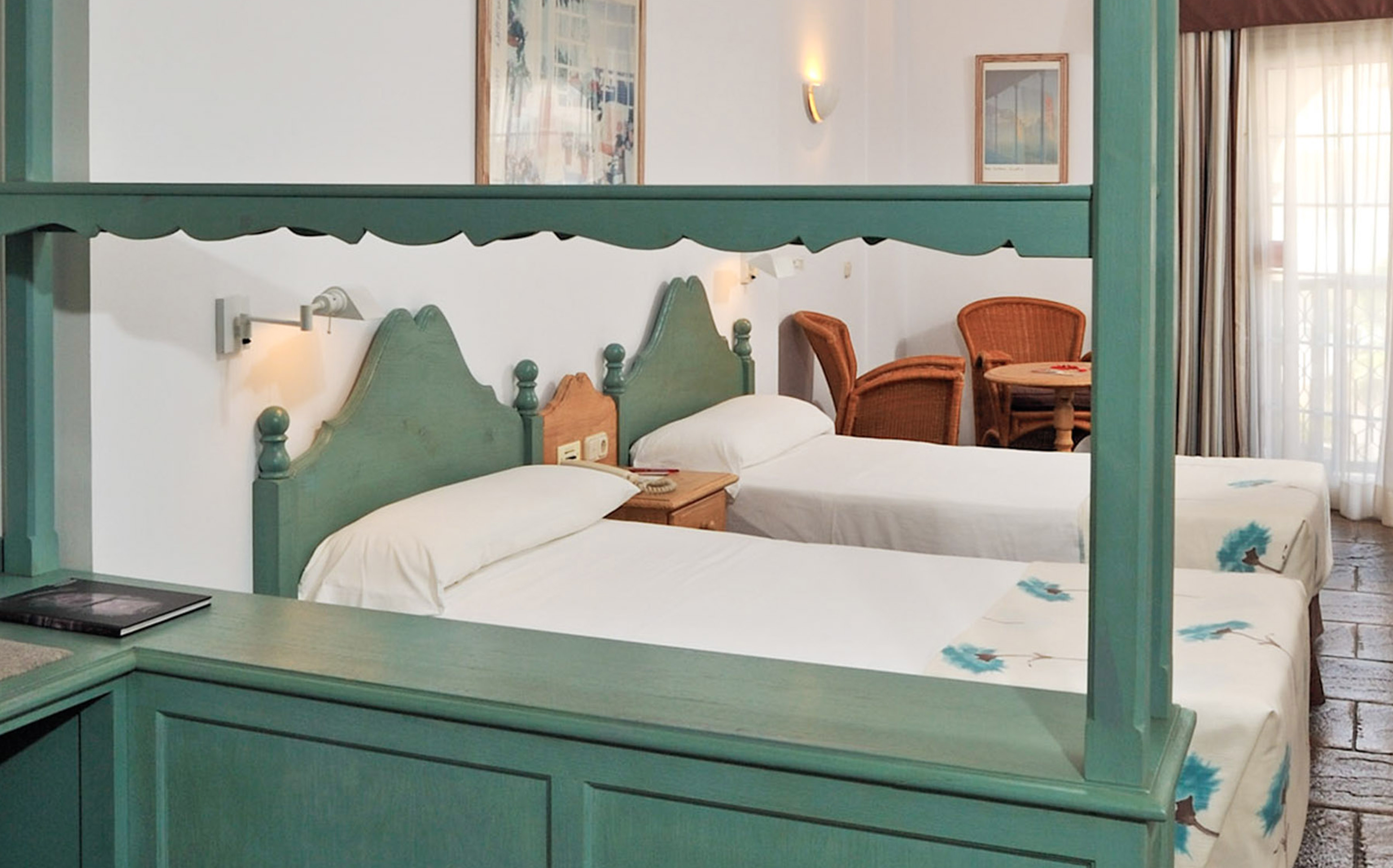 Traditional hotel room with two green twin beds, nightstand, and wicker seating.