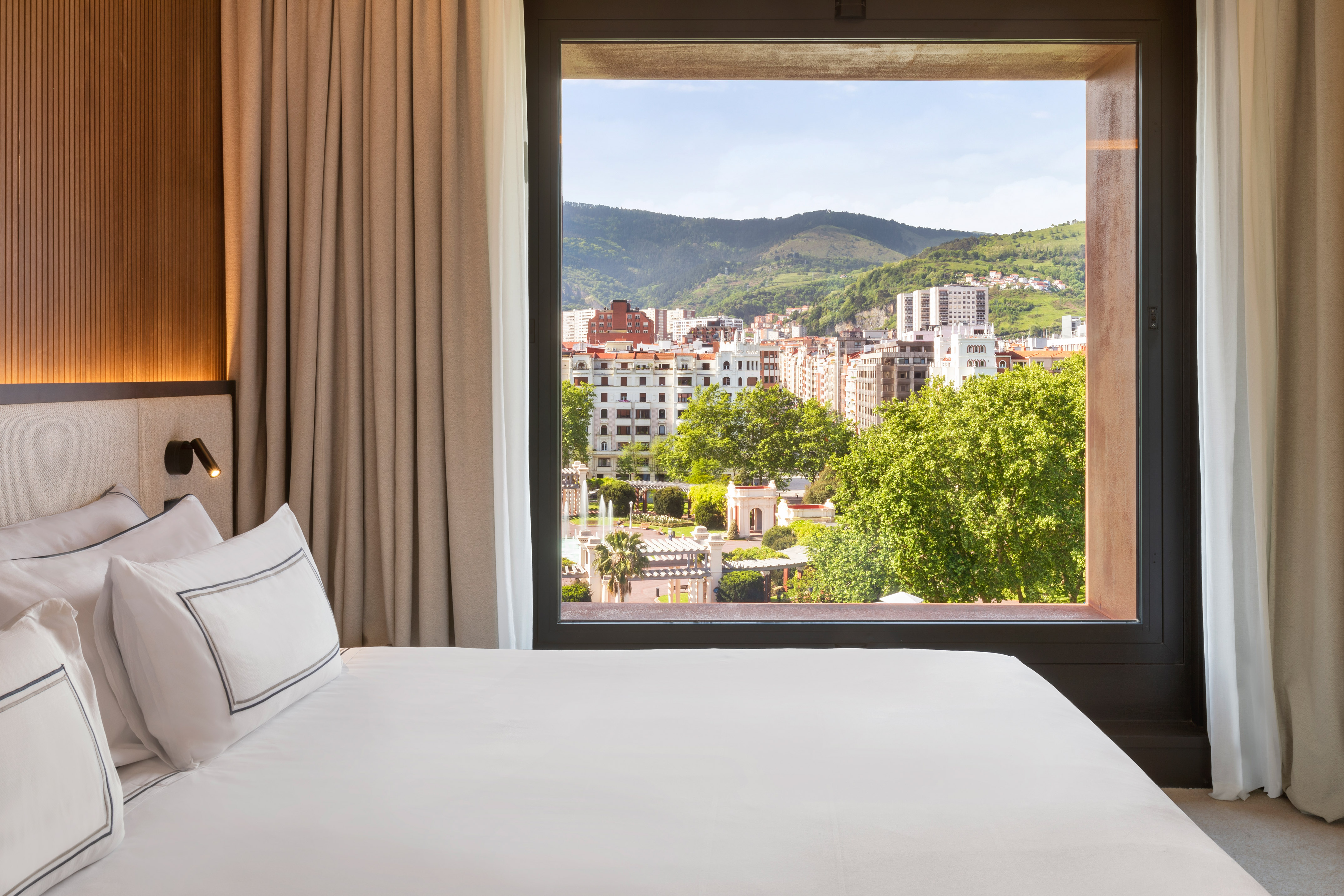 Modern hotel room bed and window view of city, park, and distant mountains.