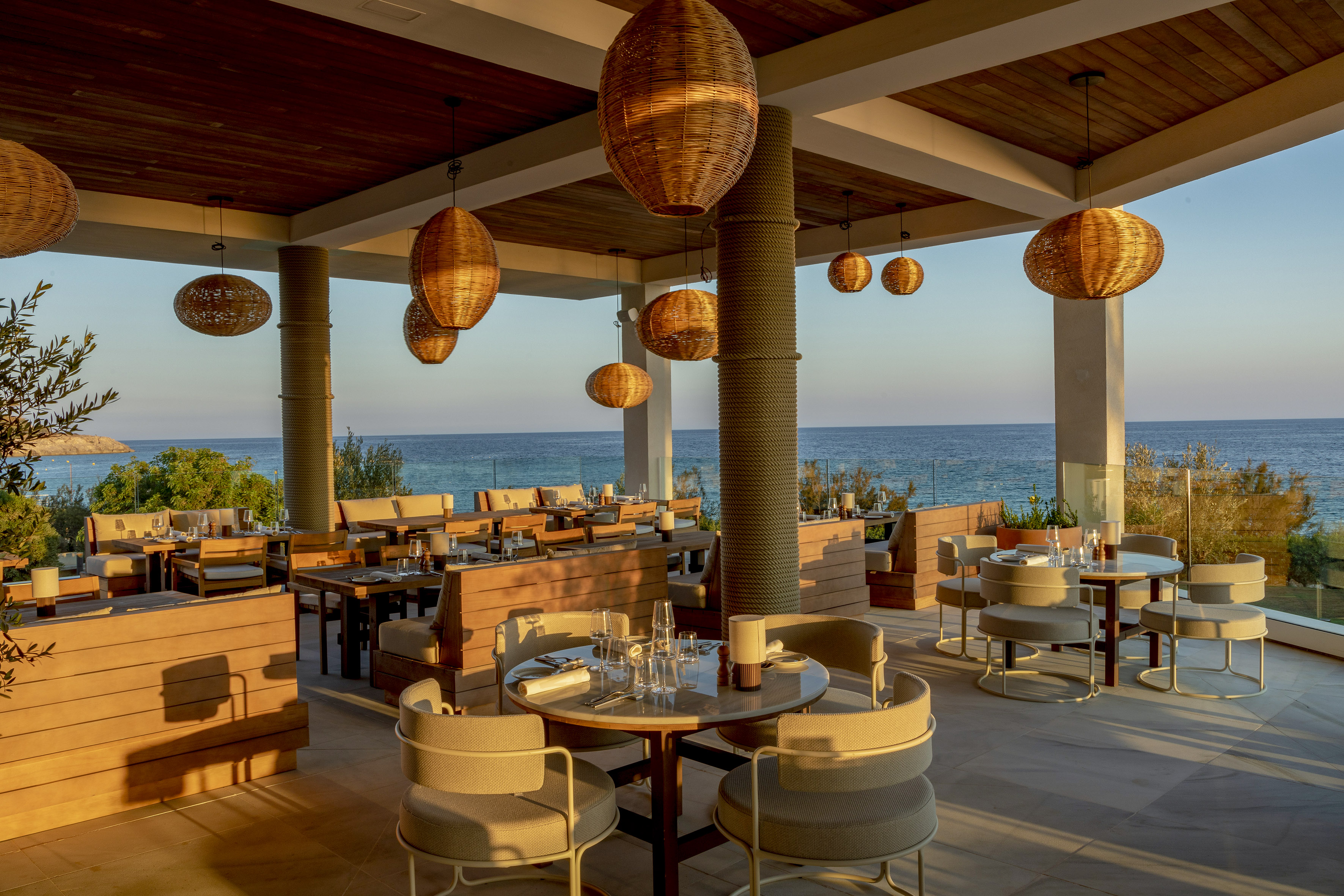 Outdoor restaurant with sea view, woven pendant lights, and modern furniture at sunset.