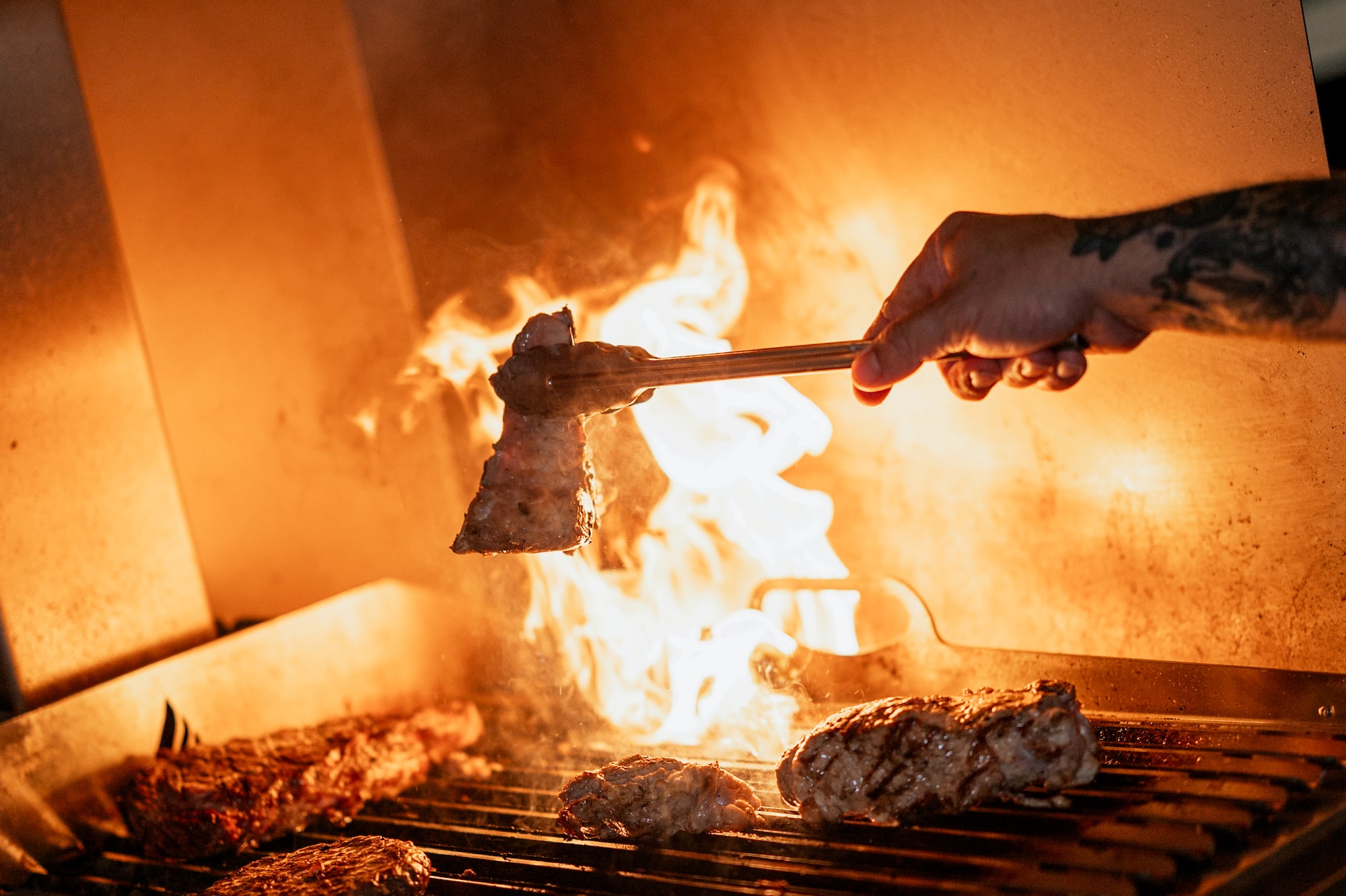 Grilling meat with tongs over fiery flames on a charcoal BBQ.