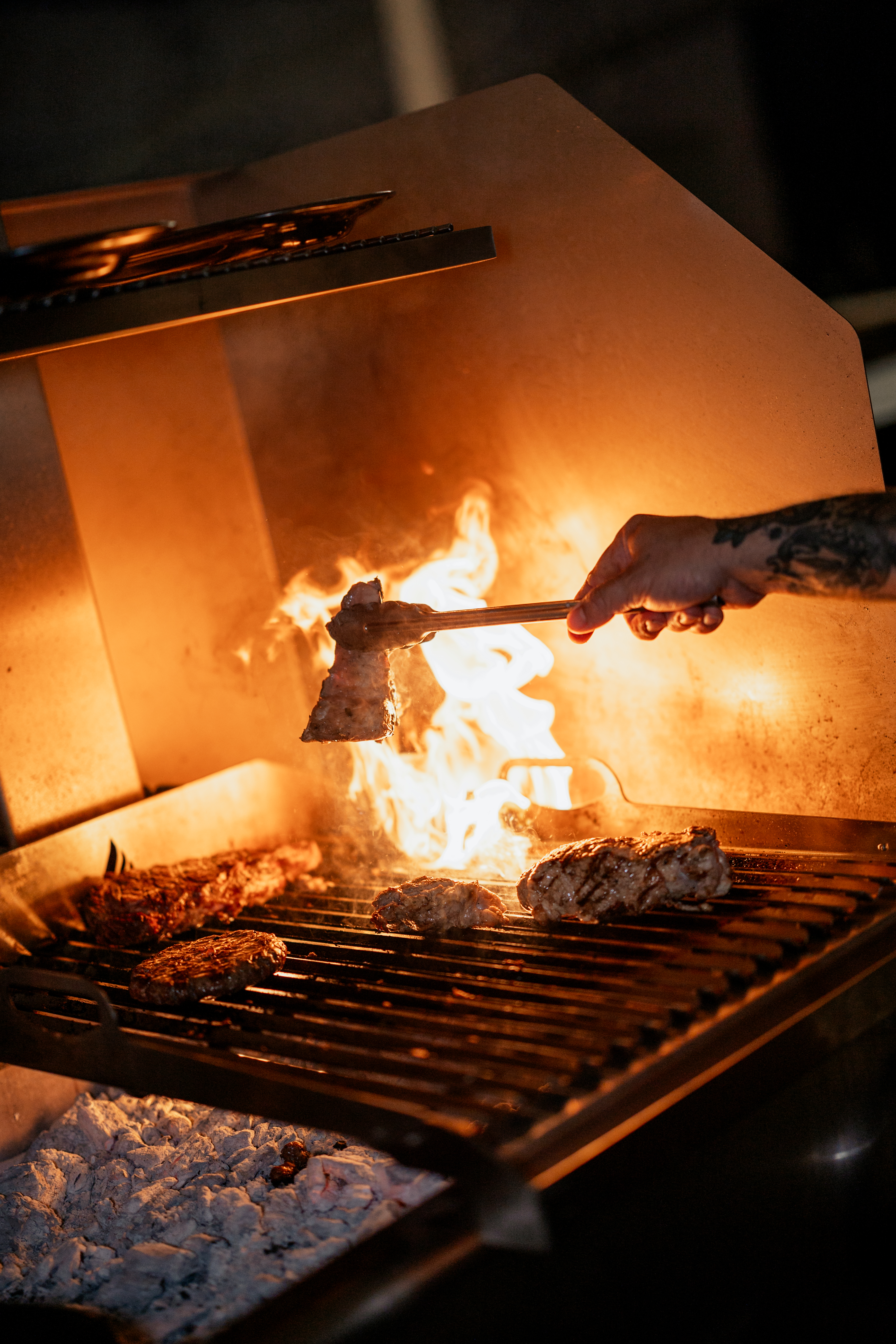 Grilling meat with tongs over fiery flames on a charcoal BBQ.