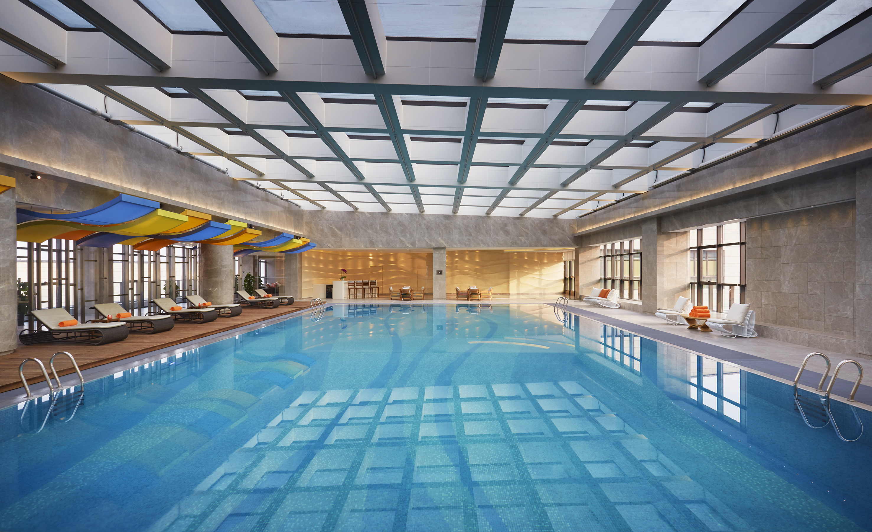 Modern indoor hotel swimming pool with skylight and poolside lounge chairs.