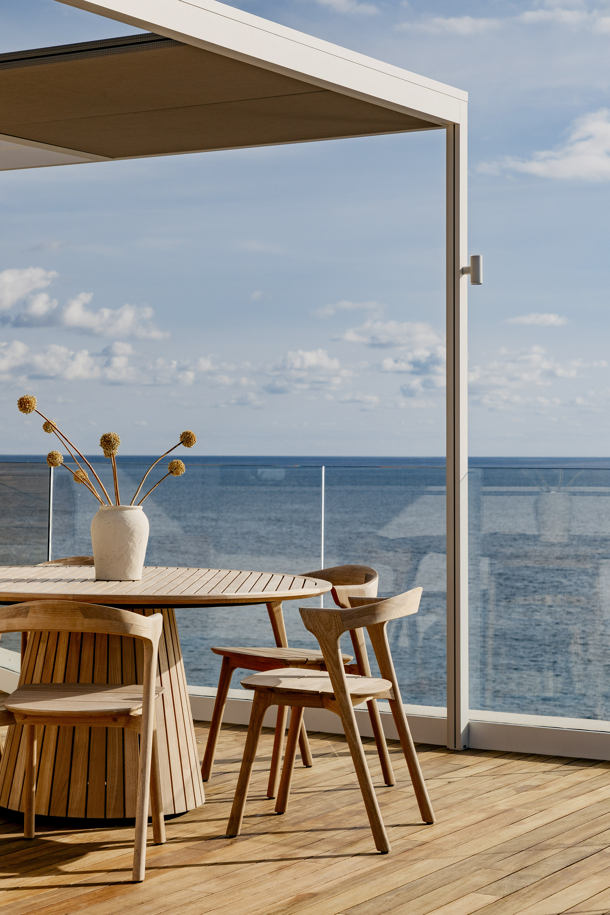 Modern wooden outdoor dining set on deck overlooking sea with pergola.