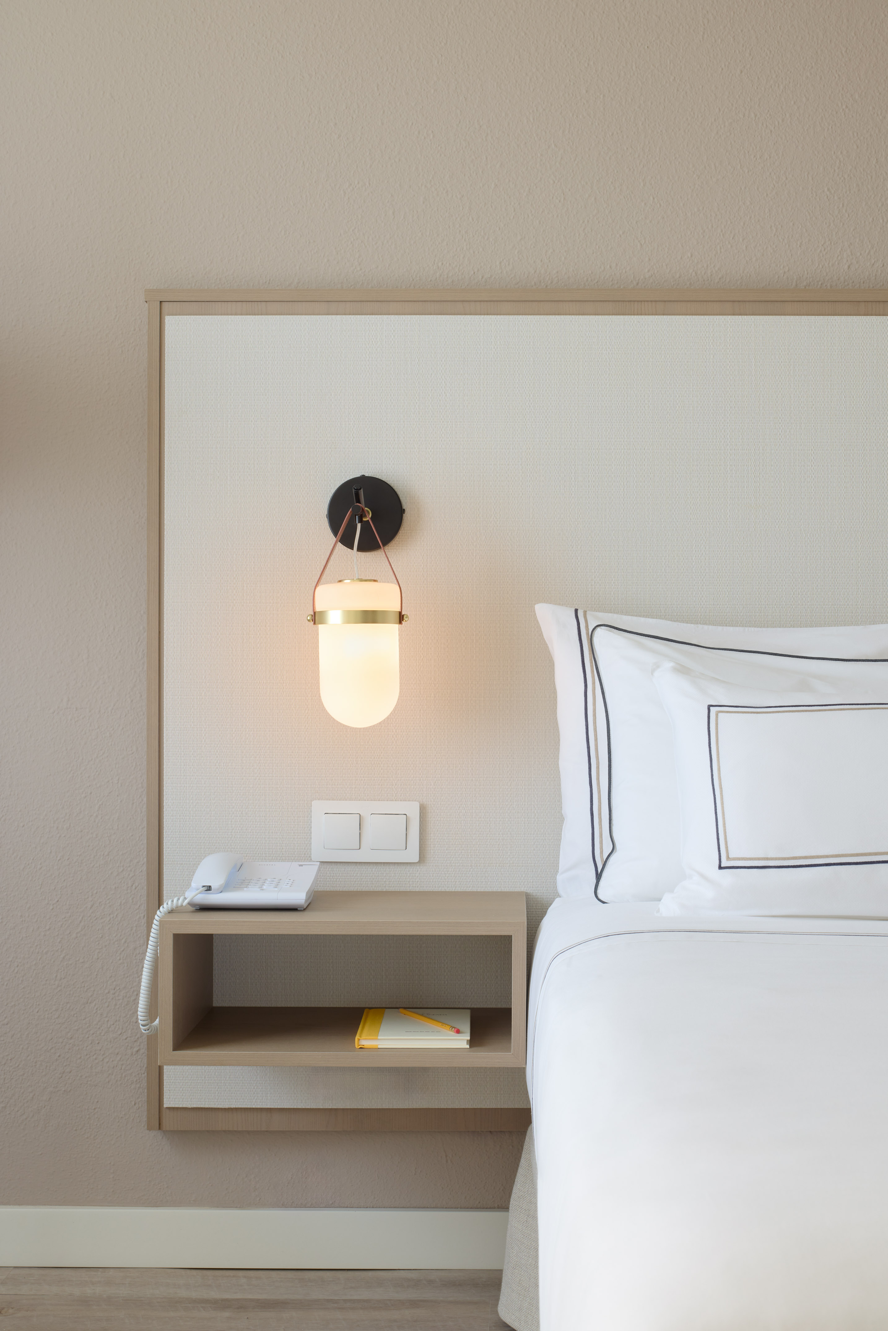 Hotel bedroom with modern bedside lamp, white bedding, and wood nightstand.
