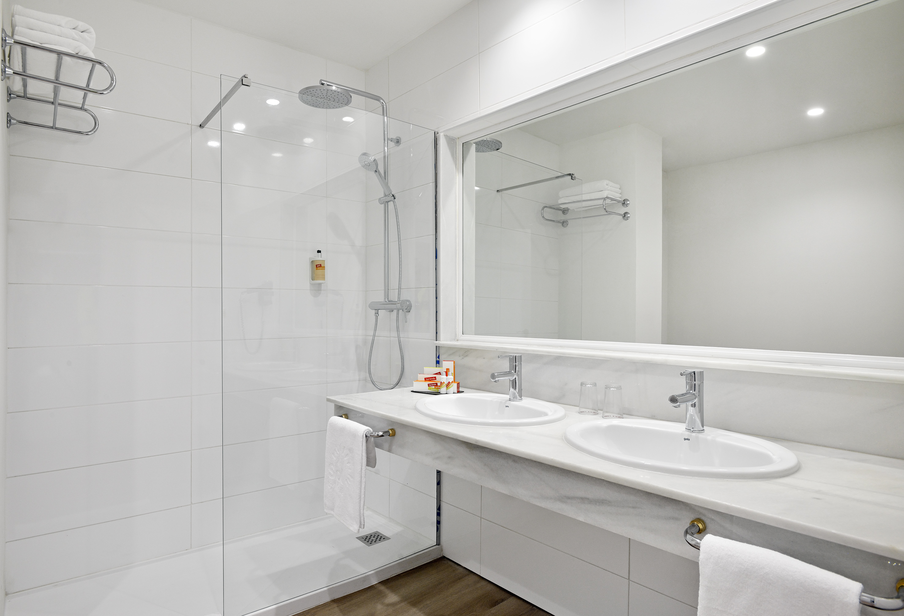 White tiled modern bathroom featuring double sinks, marble vanity, and glass shower.