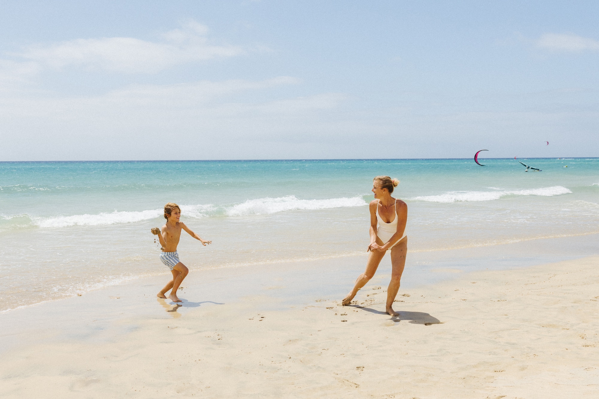 Madre e hijo jugando con arena en la playa. Mar azul, kitesurf de fondo. Vacaciones familiares.