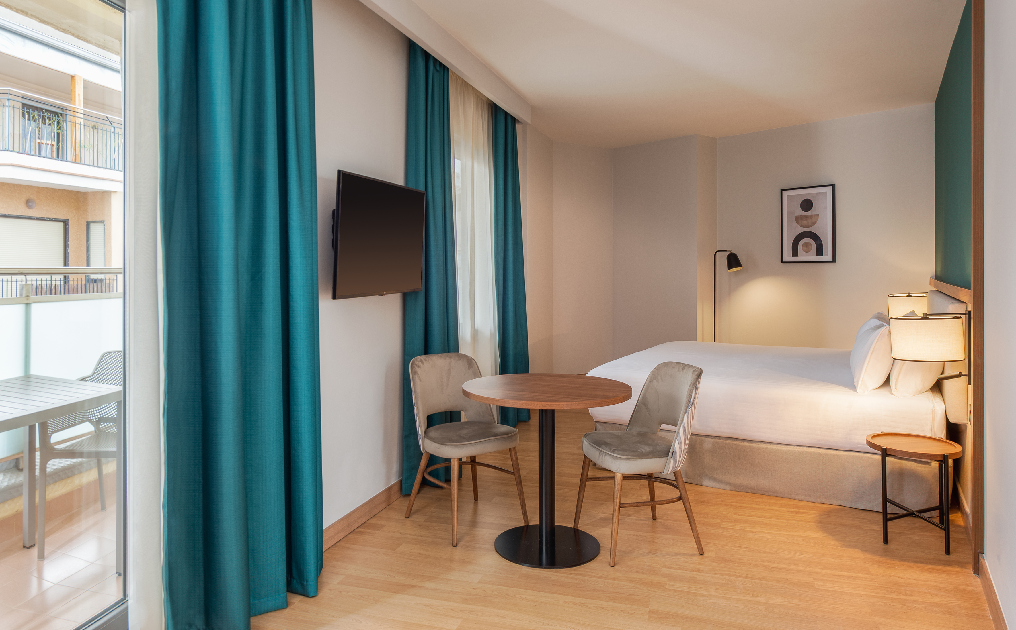 Modern hotel suite with bed, small table, two chairs, TV, and balcony access.