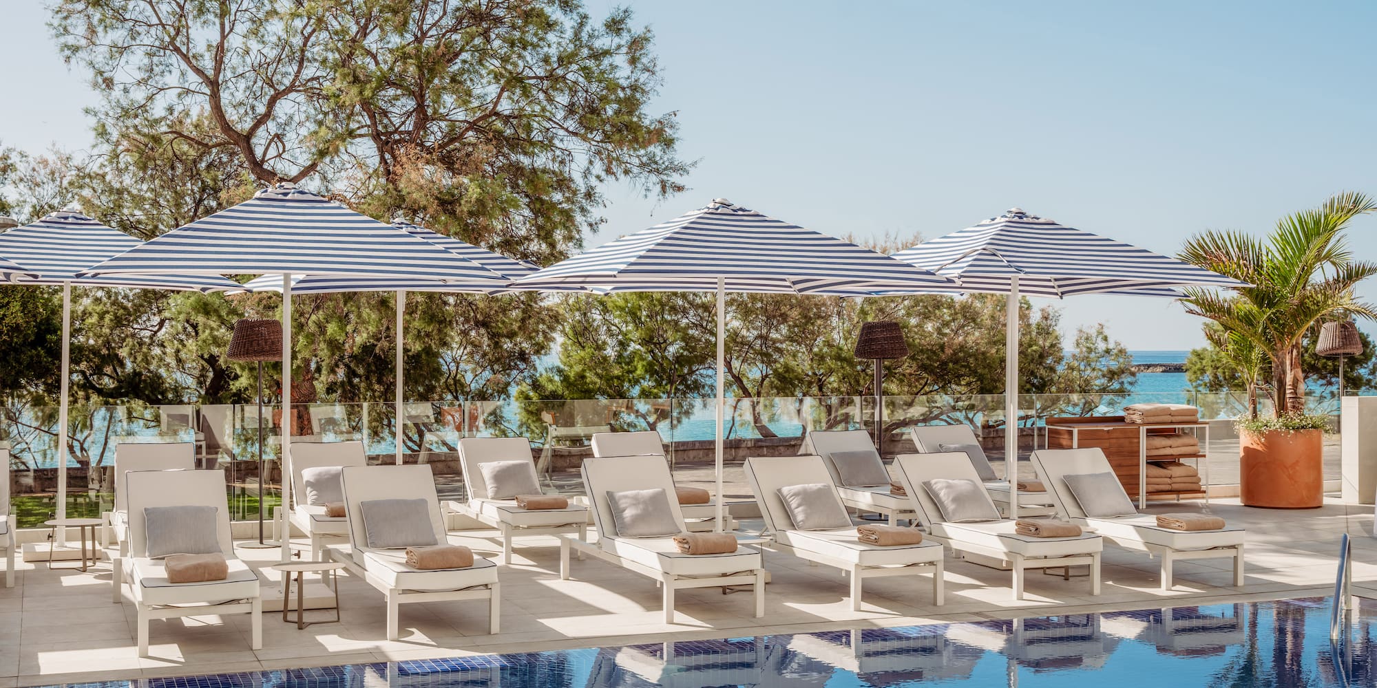 Piscina de resort con tumbonas blancas y sombrillas a rayas azules, vista al mar.