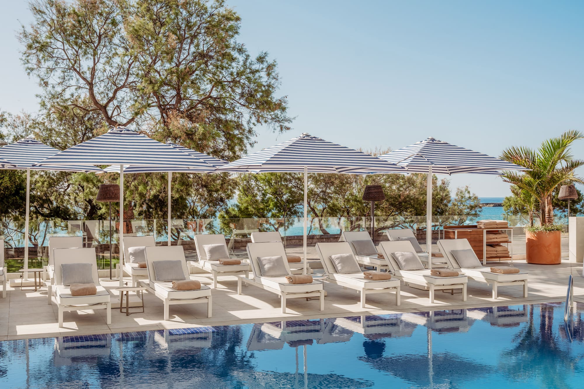 Piscina de resort con tumbonas blancas y sombrillas a rayas azules, vista al mar.