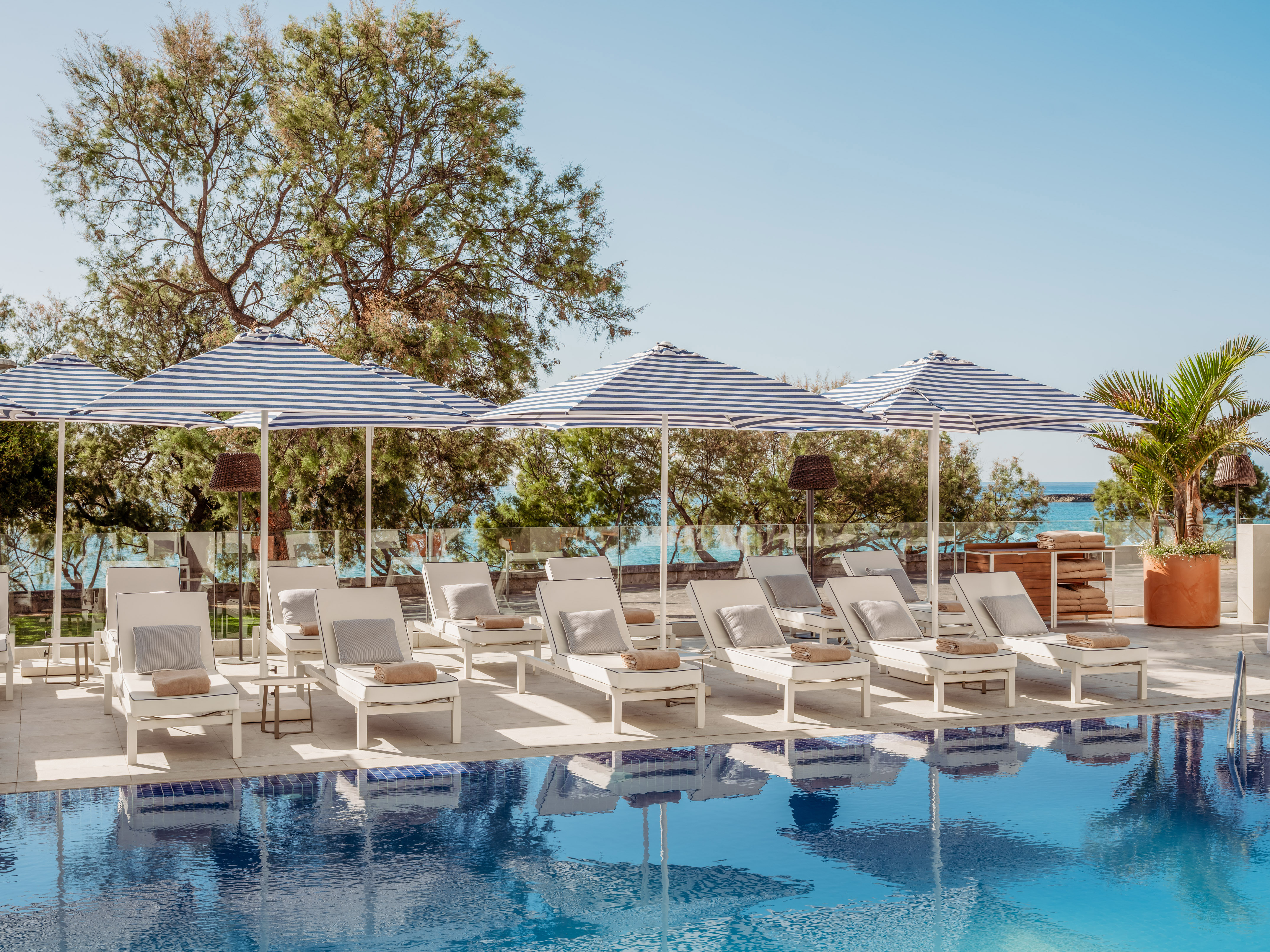 Piscina de resort con tumbonas blancas y sombrillas a rayas azules, vista al mar.