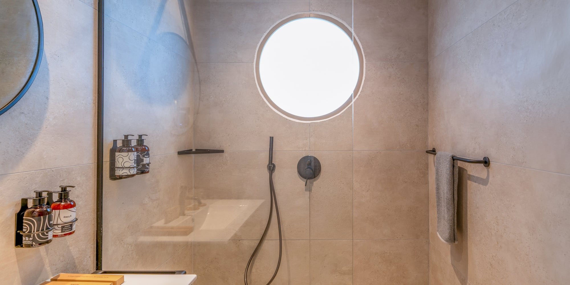 Beige tiled modern shower with glass partition, rainfall head, and circular window.