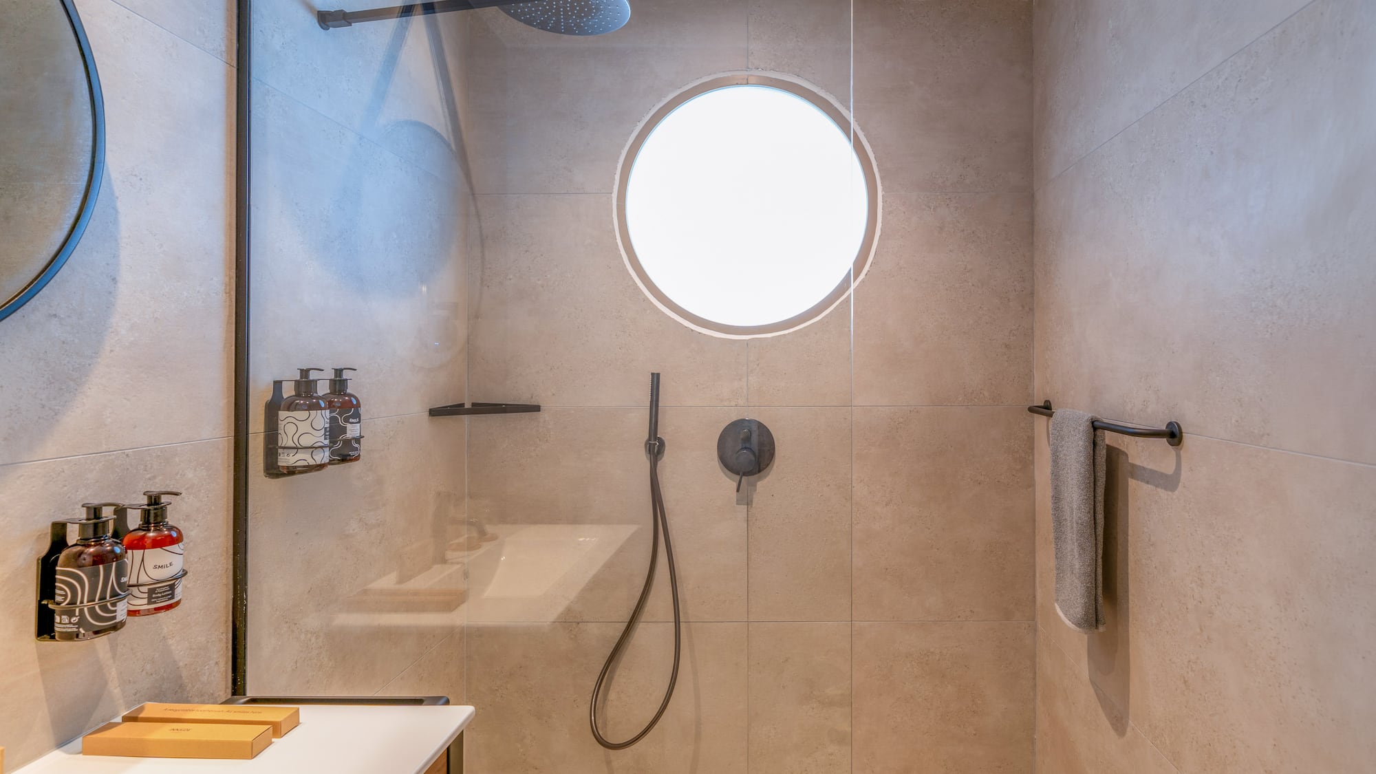 Beige tiled modern shower with glass partition, rainfall head, and circular window.