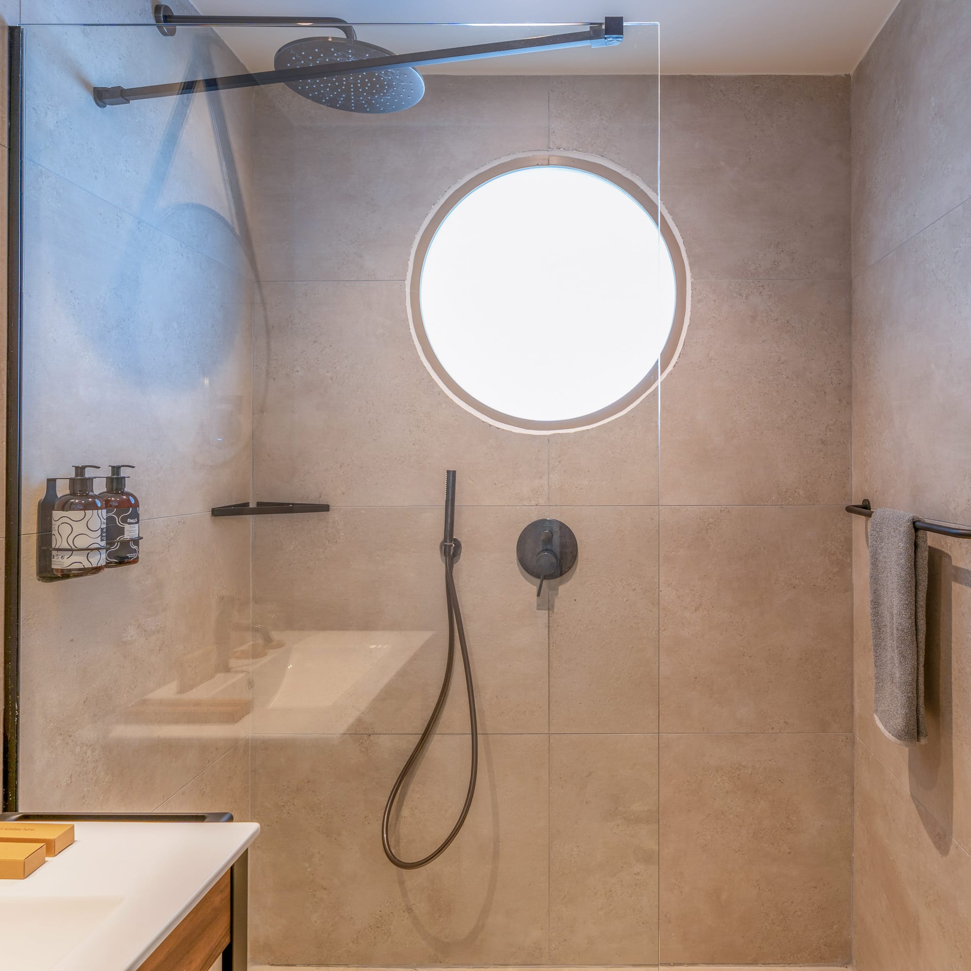 Beige tiled modern shower with glass partition, rainfall head, and circular window.