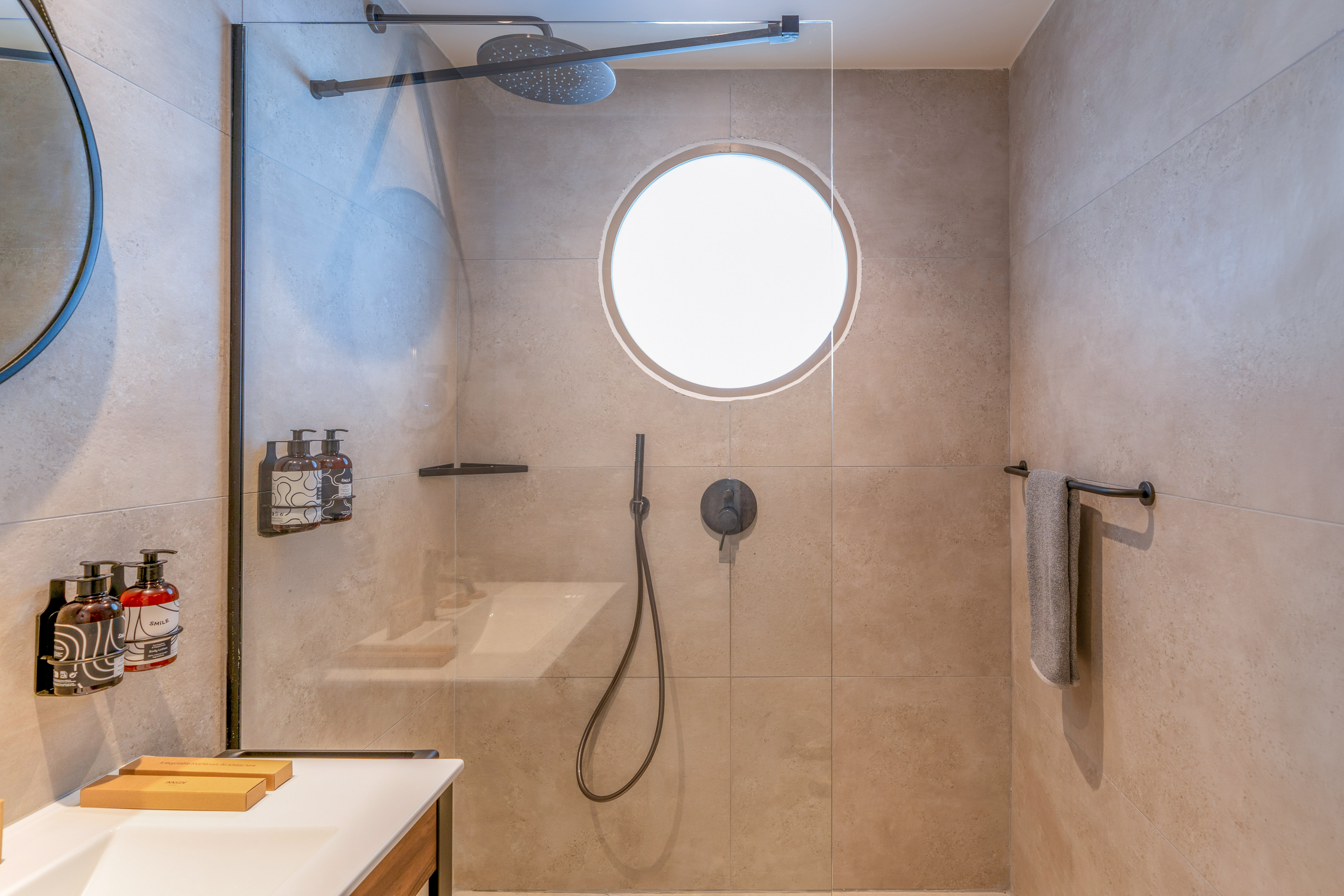 Beige tiled modern shower with glass partition, rainfall head, and circular window.