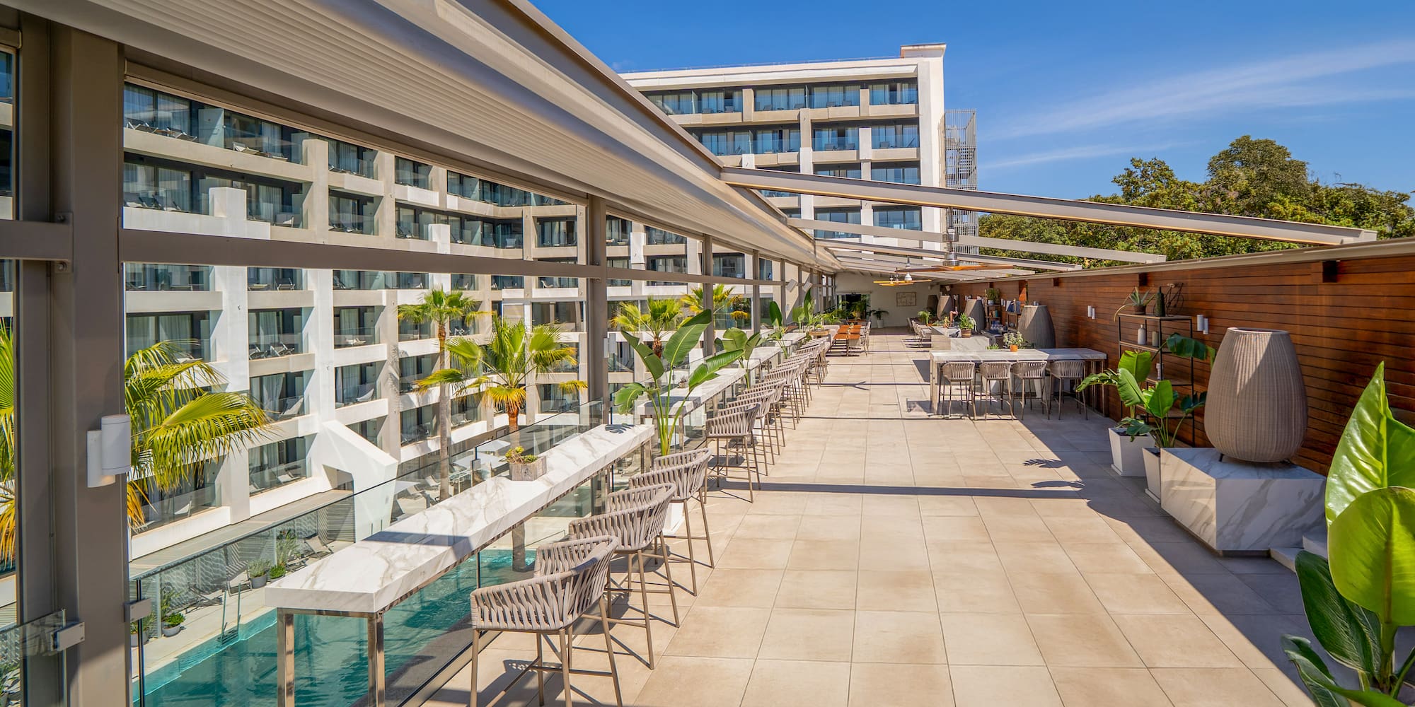Modern rooftop bar and patio overlooking a hotel pool with palm trees.
