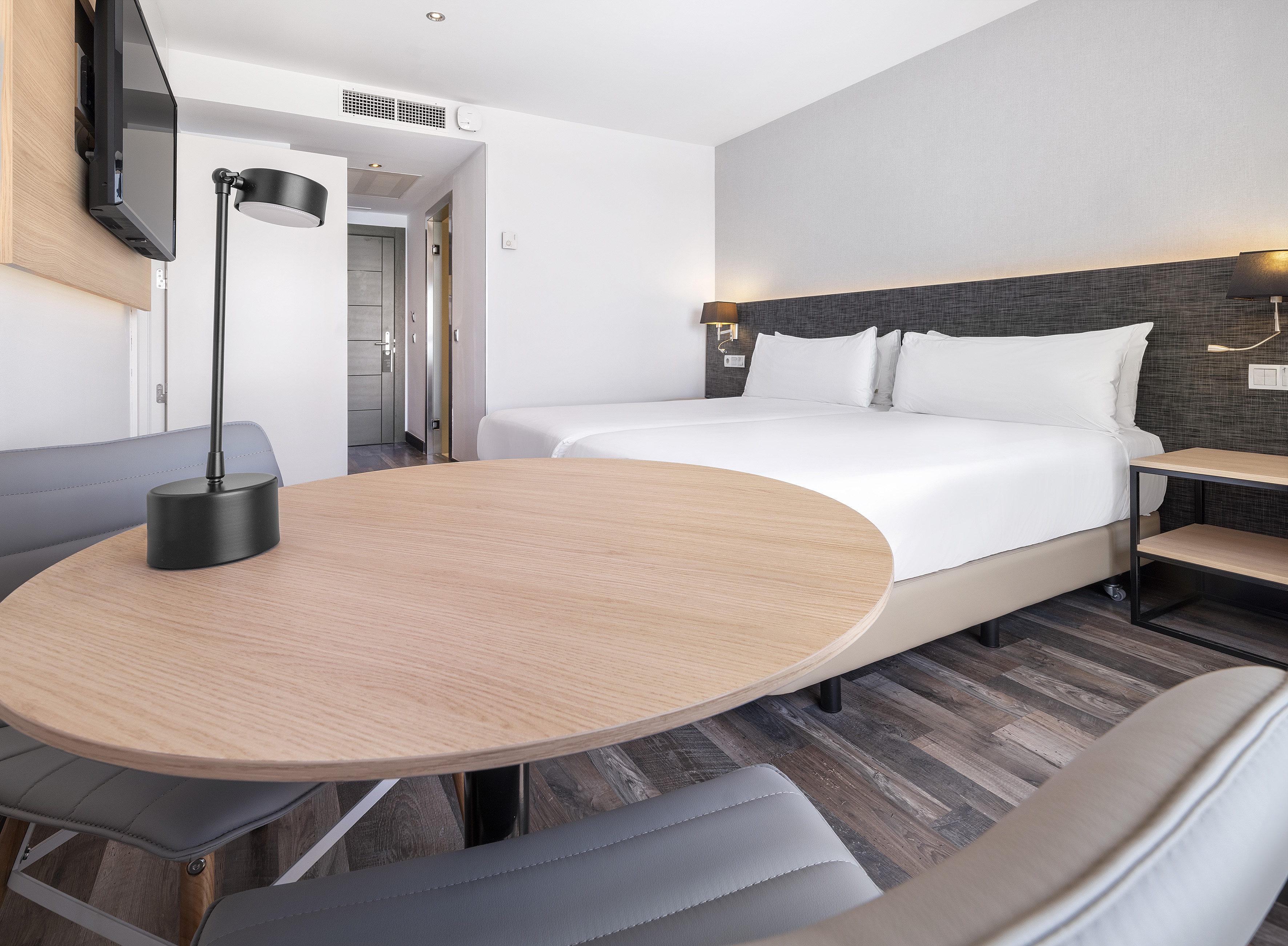 Modern hotel room featuring twin beds, a round wooden table, and sleek chairs.