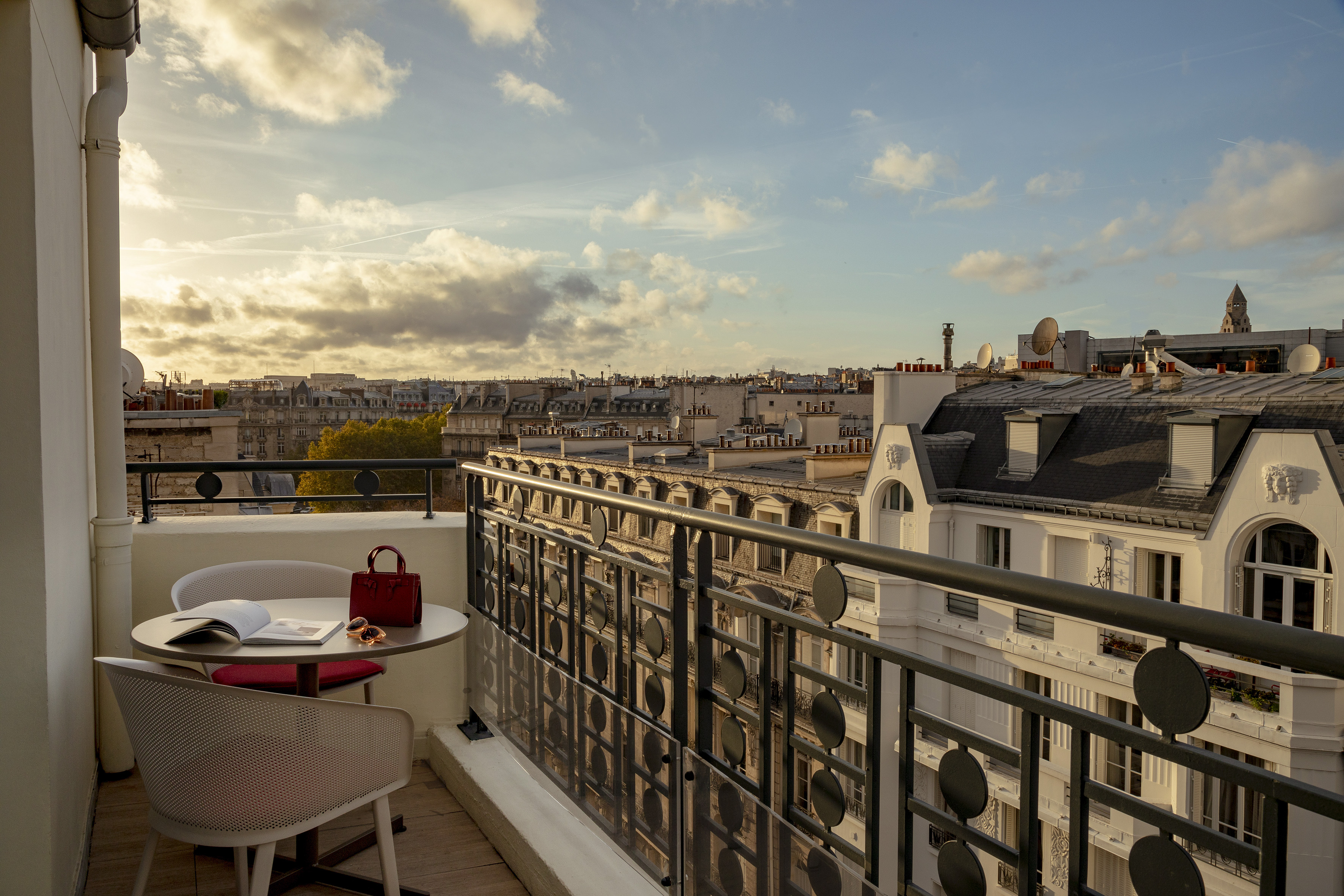 Paris balcony view with table, book, handbag, and city rooftops at sunset.
