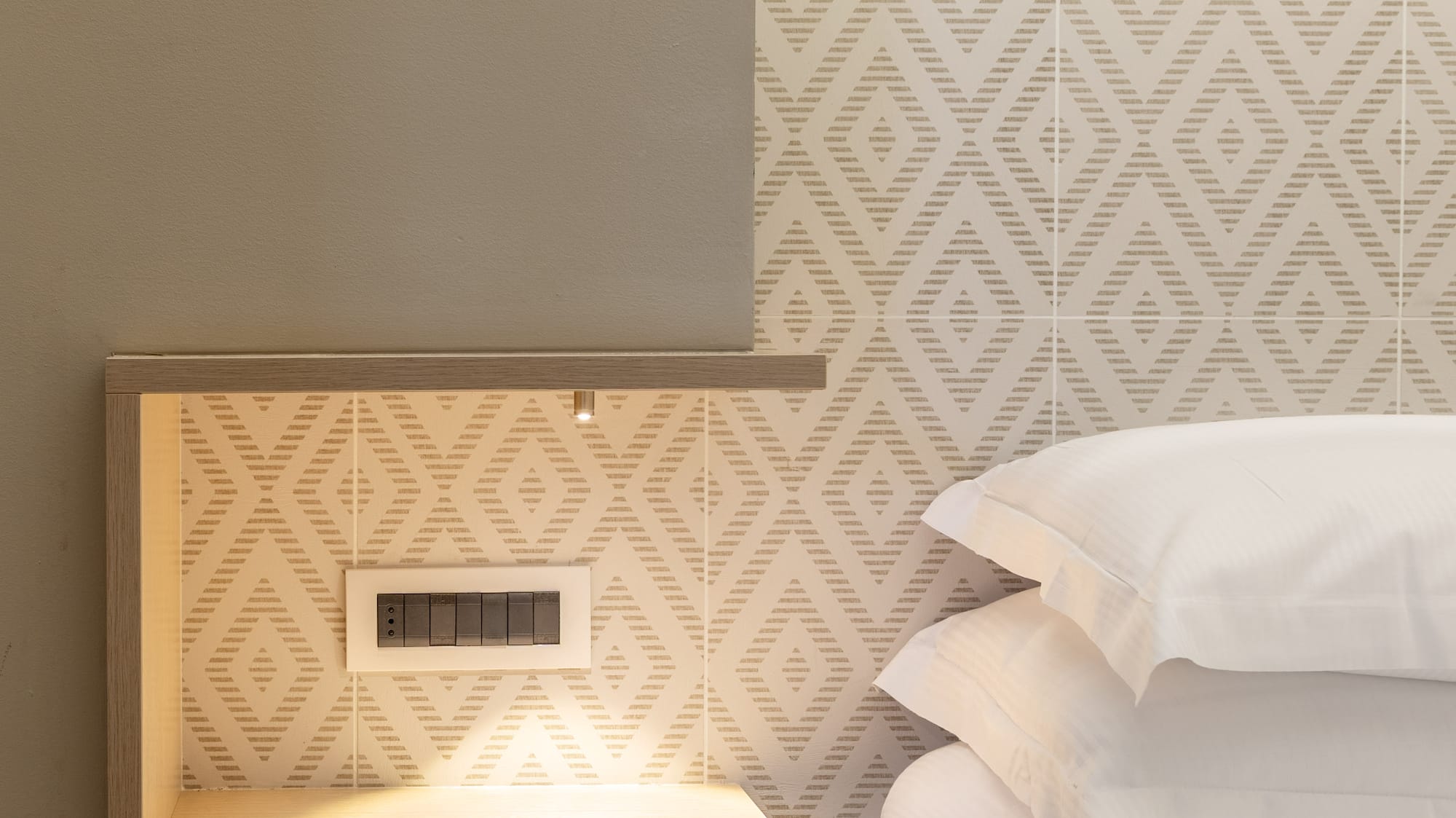 Hotel bedroom: modern nightstand with integrated light, geometric wall, white bed and pillows.