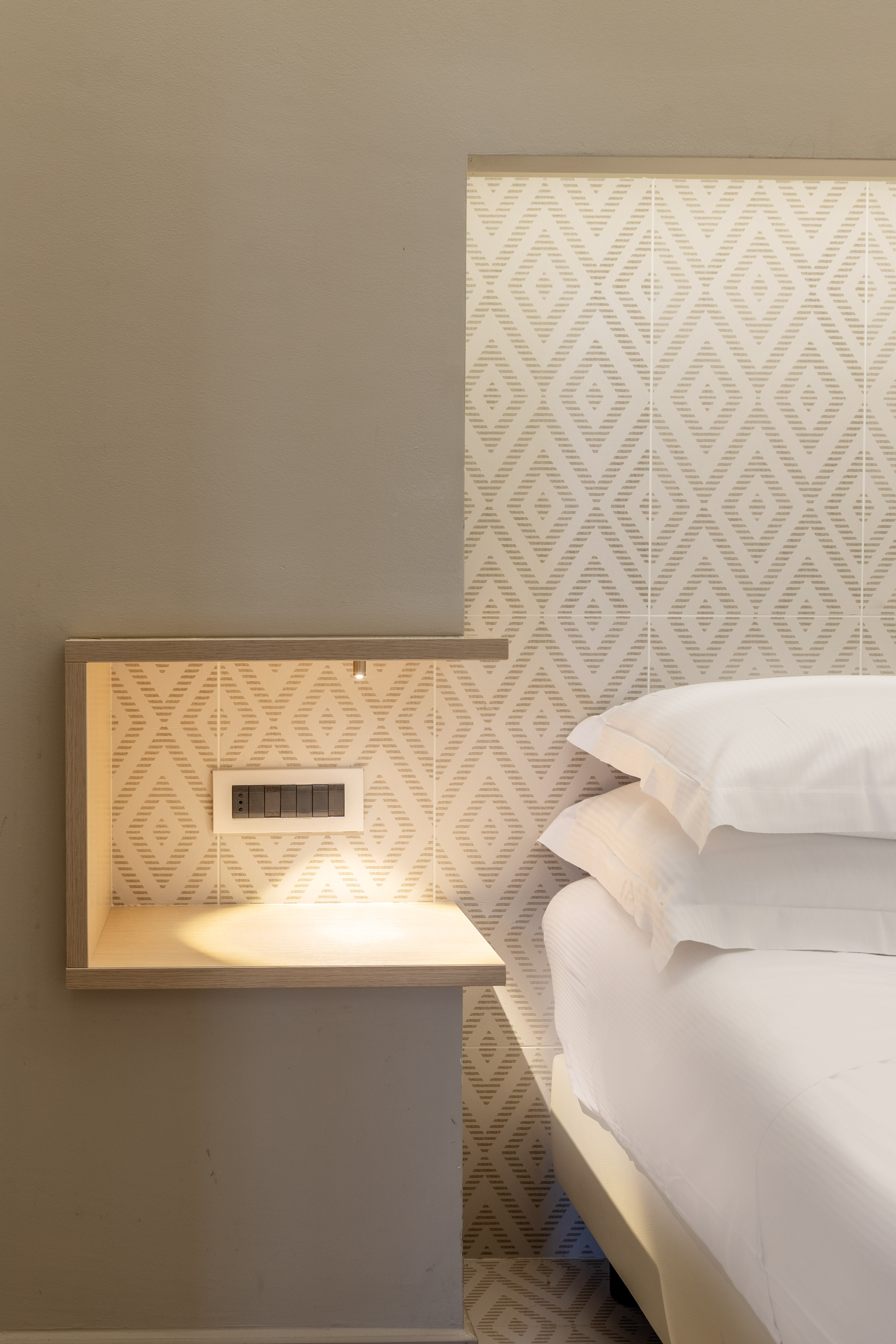 Hotel bedroom: modern nightstand with integrated light, geometric wall, white bed and pillows.