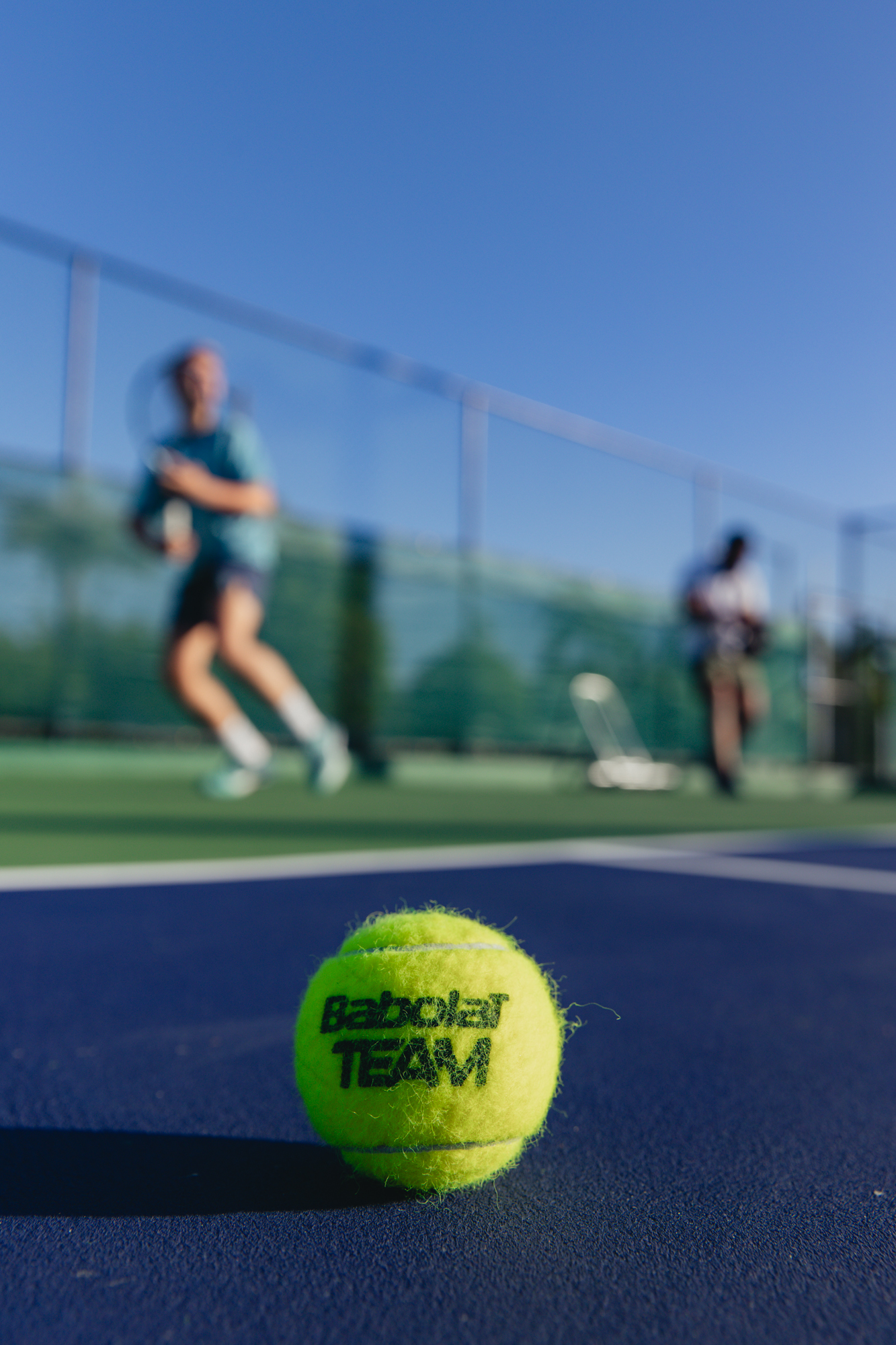 Close-up of Babolat tennis ball on blue court, players training in background.