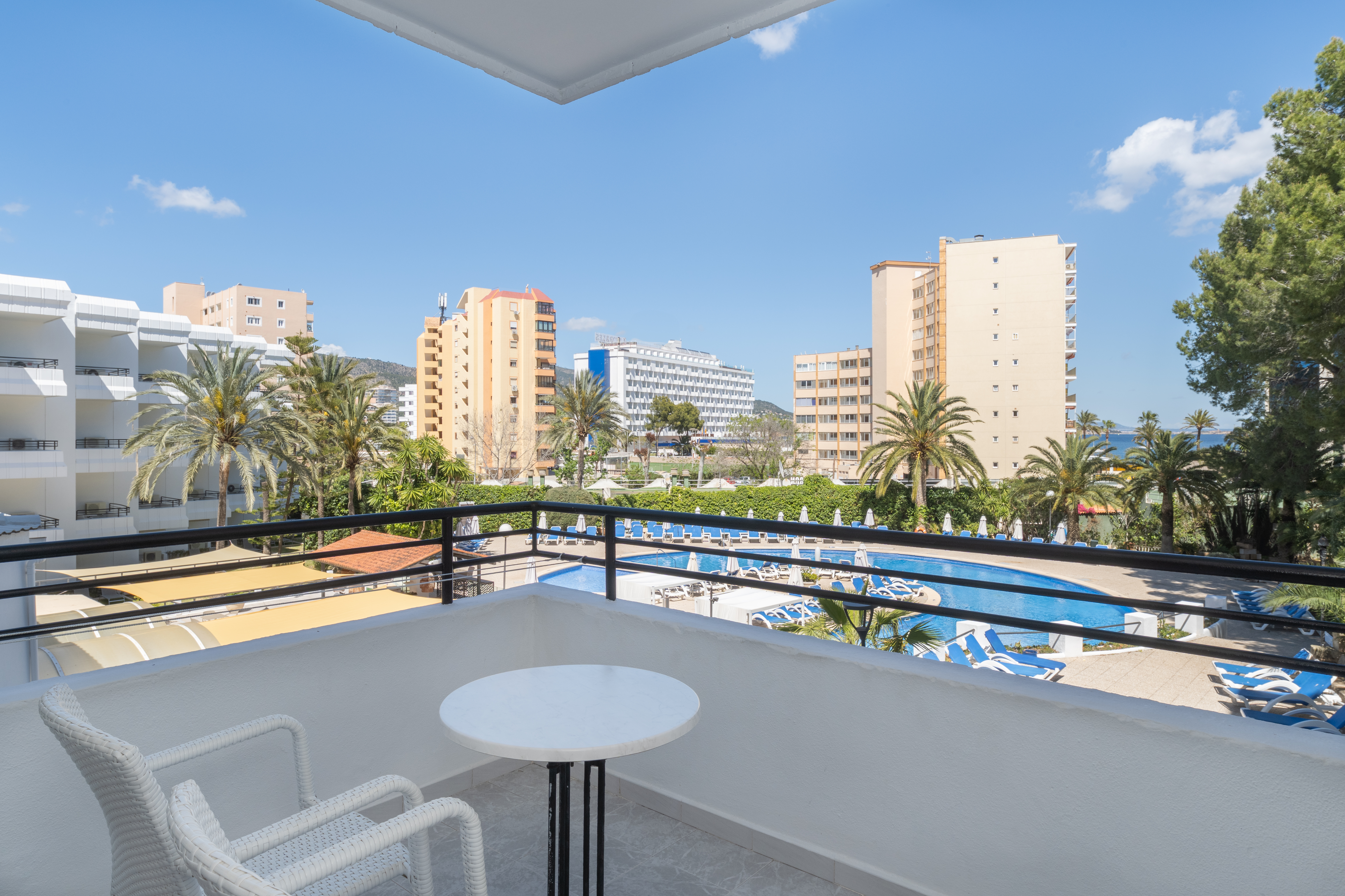 Balcony overlooking hotel pool, palm trees, resort buildings, and sea.