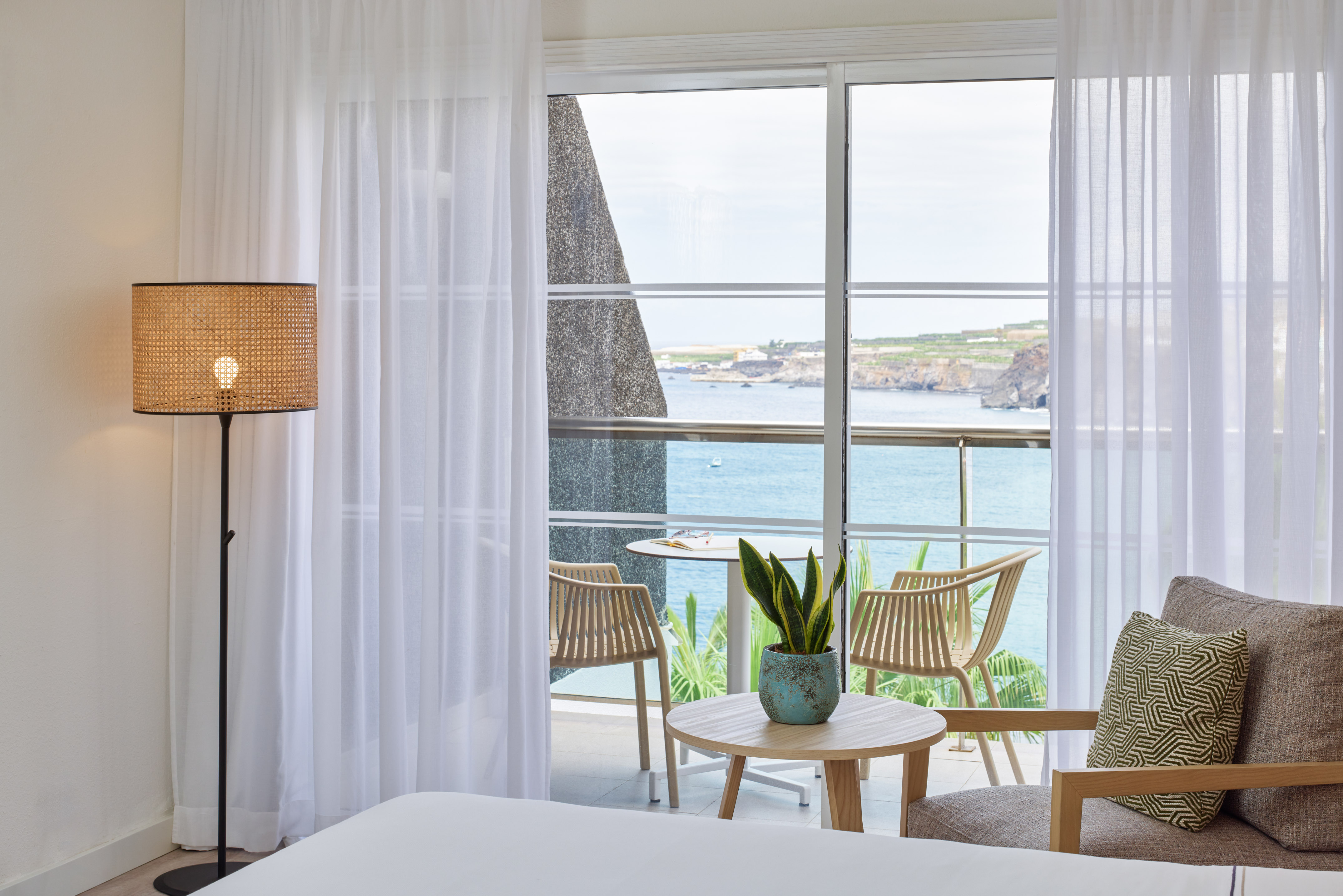 Modern hotel room with ocean view balcony, white curtains, and rattan lamp.
