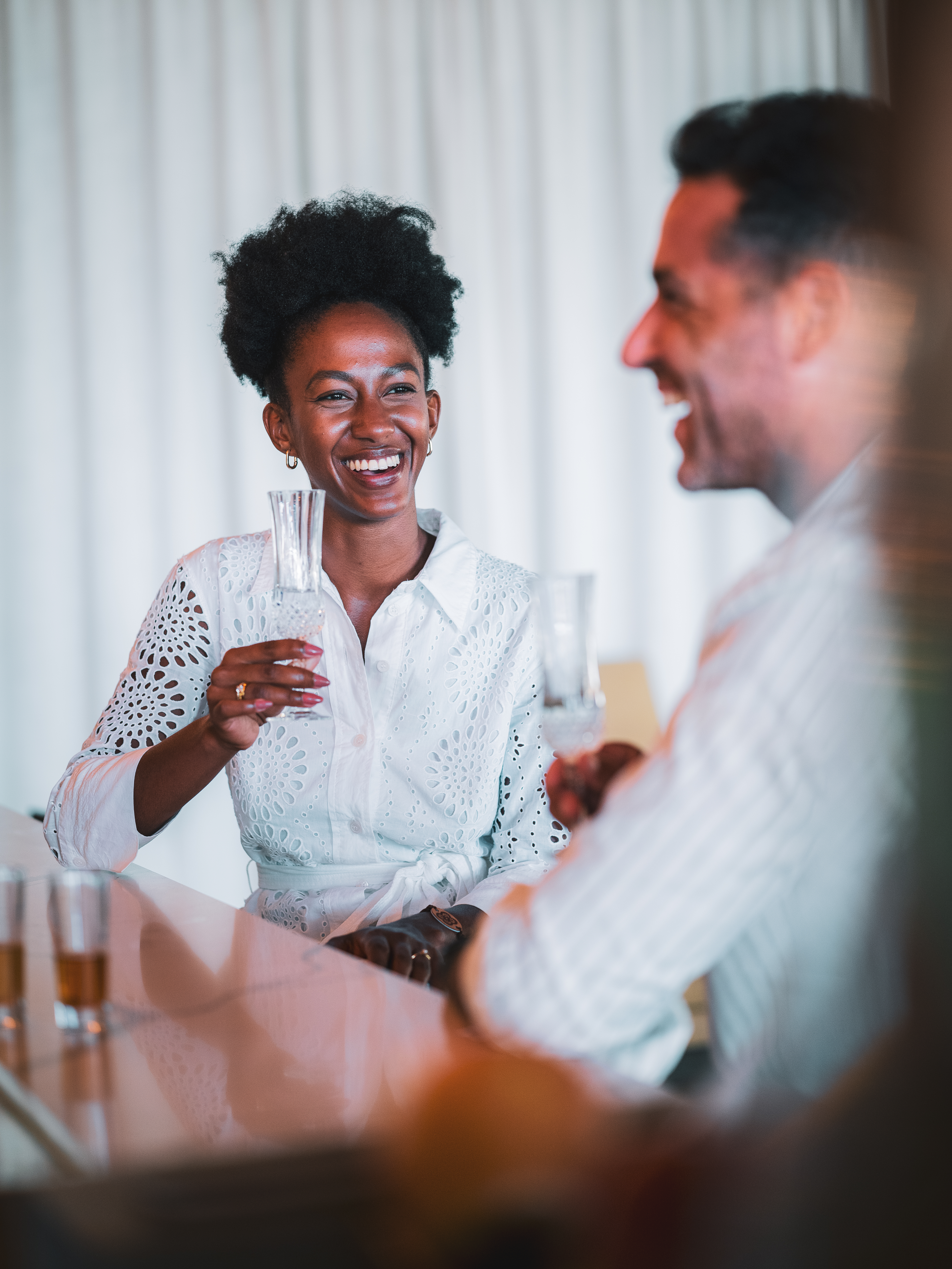 Smiling Black woman in white shirt with drink, conversing happily with man at bar.