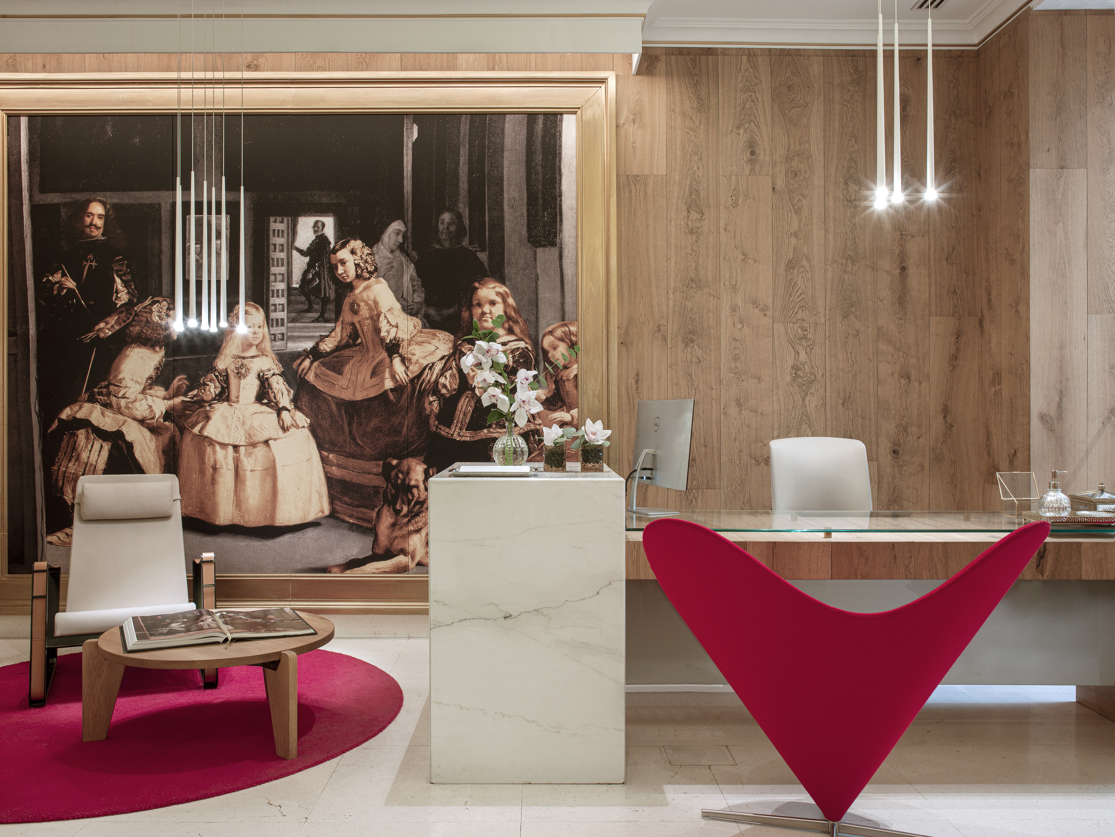 Hotel lobby with 'Las Meninas' artwork, modern reception desk, and red lounge chair.