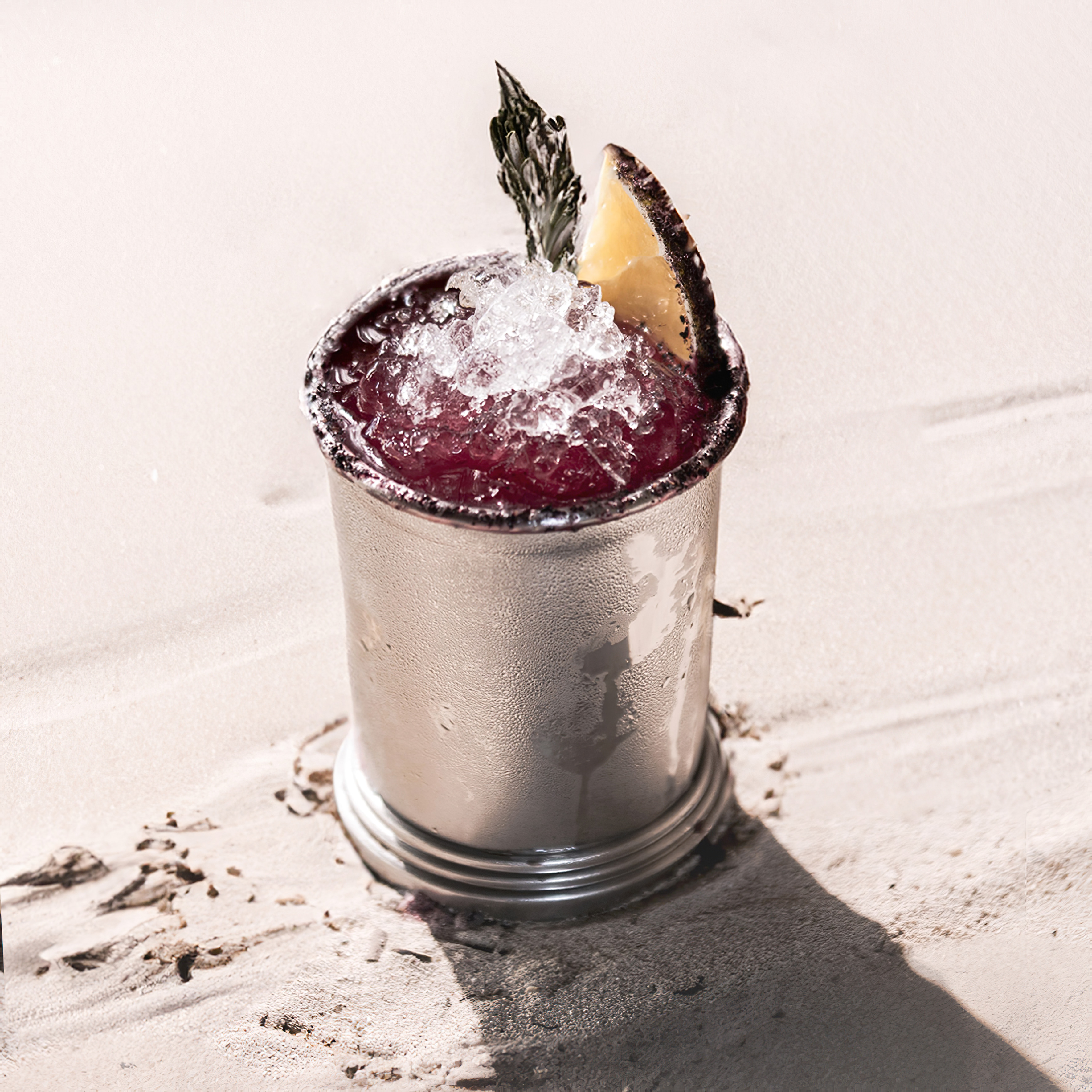 Red cocktail in silver julep cup with crushed ice, citrus, and herb on sand.