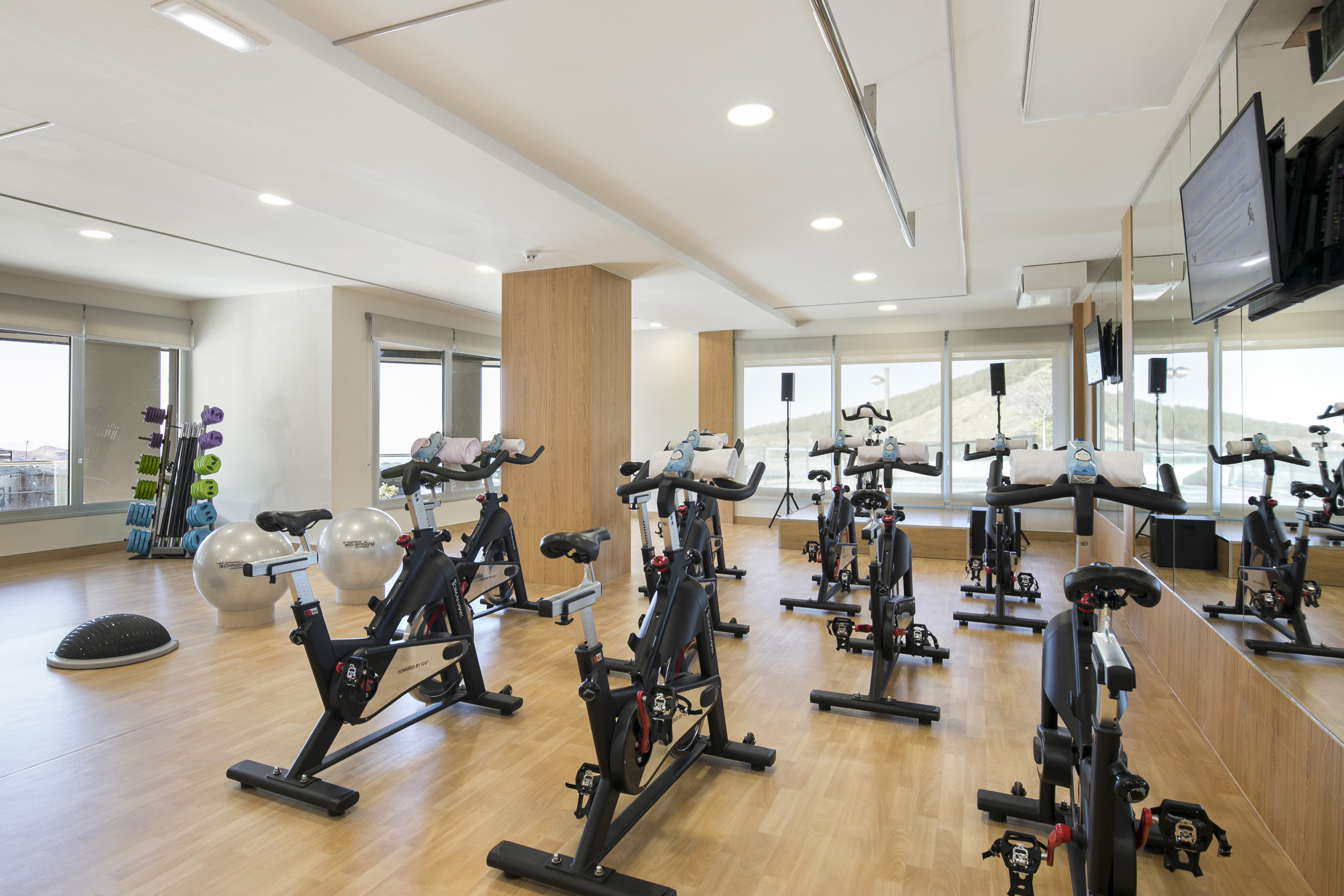 Gym with spin bikes, exercise equipment, wooden floor, and large windows.