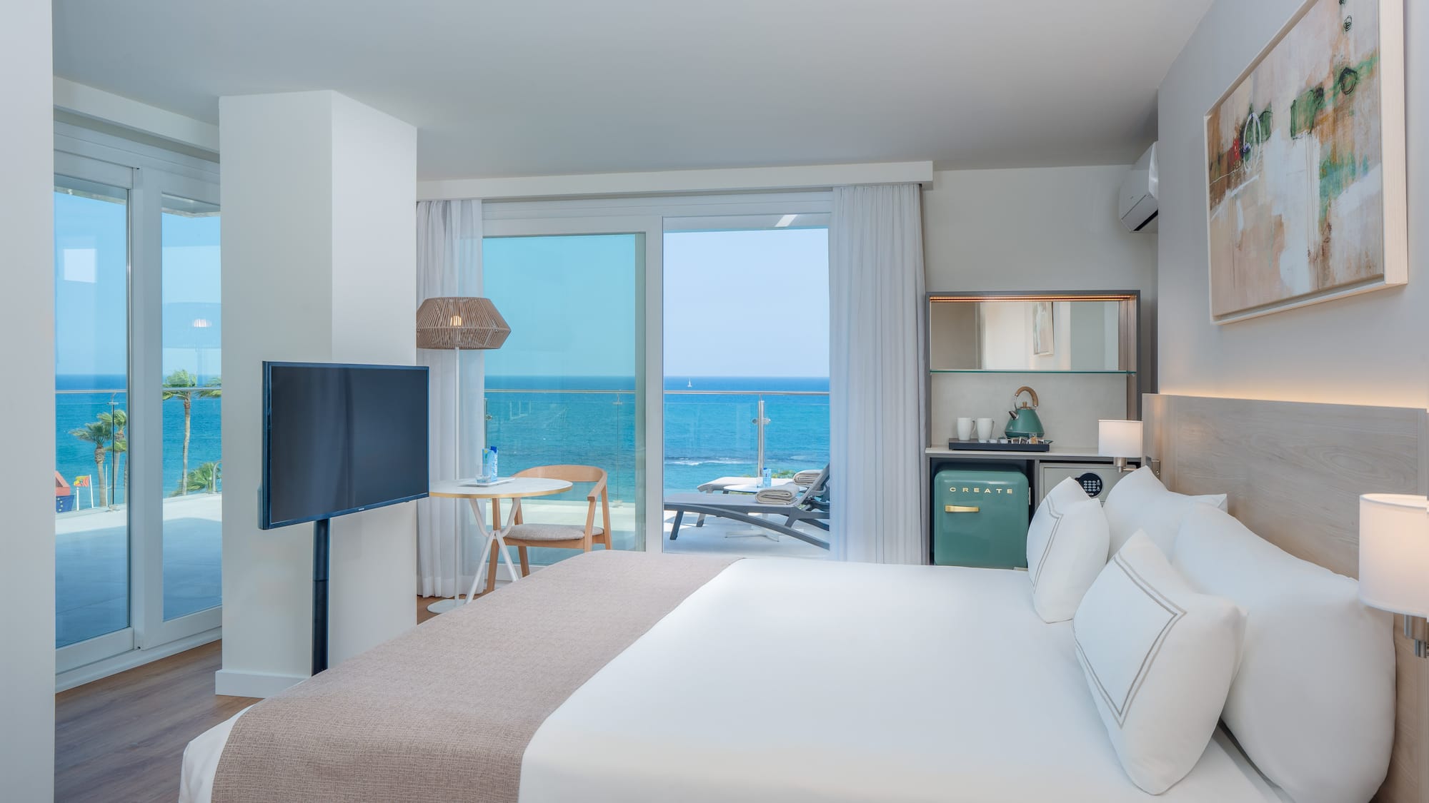 Modern hotel room with ocean view balcony, large bed, TV, and mini-bar.